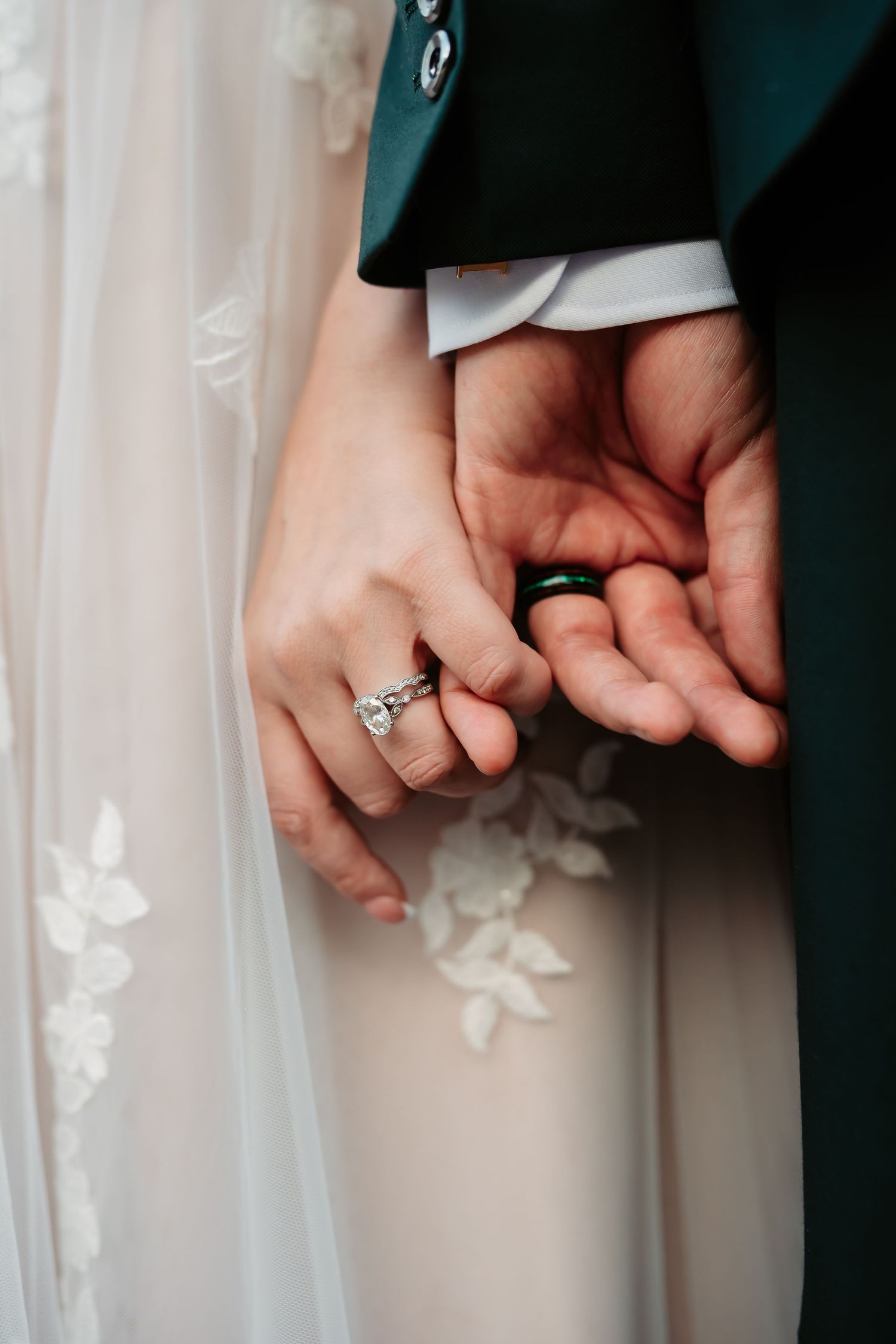 A close-up of a couple holding hands with an engagement ring visible against a white lace dress background.