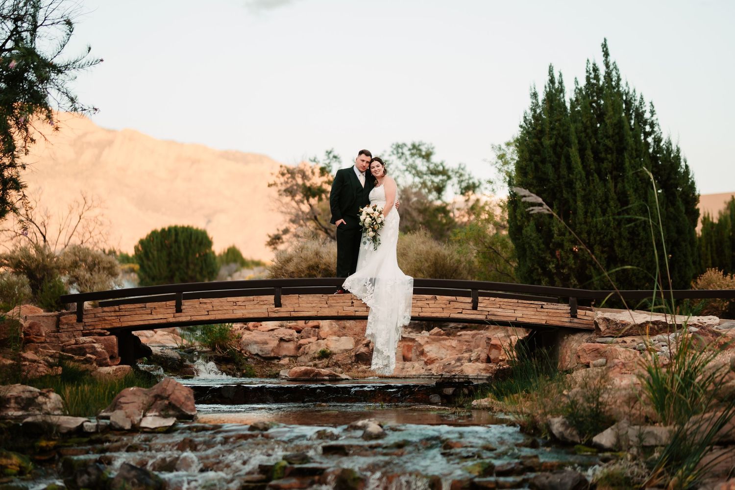 A couple poses on a rustic wooden bridge over a stream during sunset in a romantic outdoor setting.