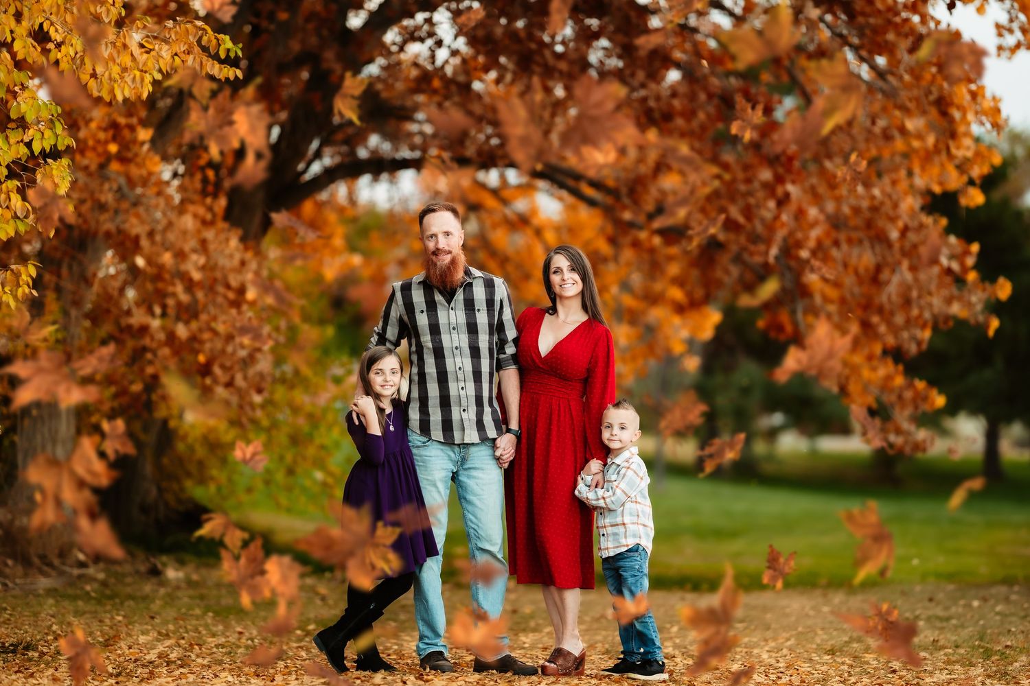 A family stands together under vibrant orange autumn foliage in a park setting during fall.