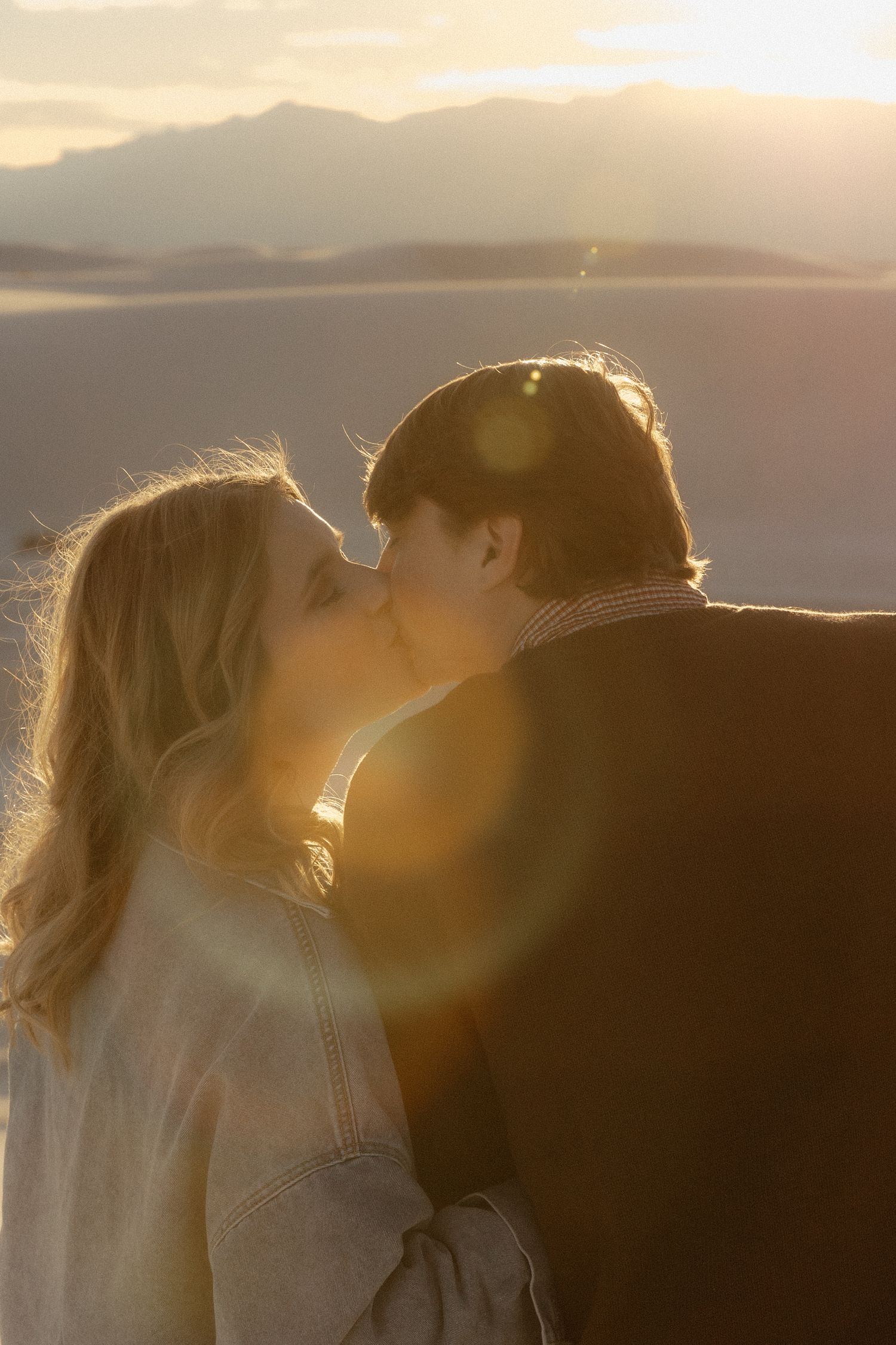 Couple sharing a romantic kiss at sunset in a desert landscape with golden light and lens flare.