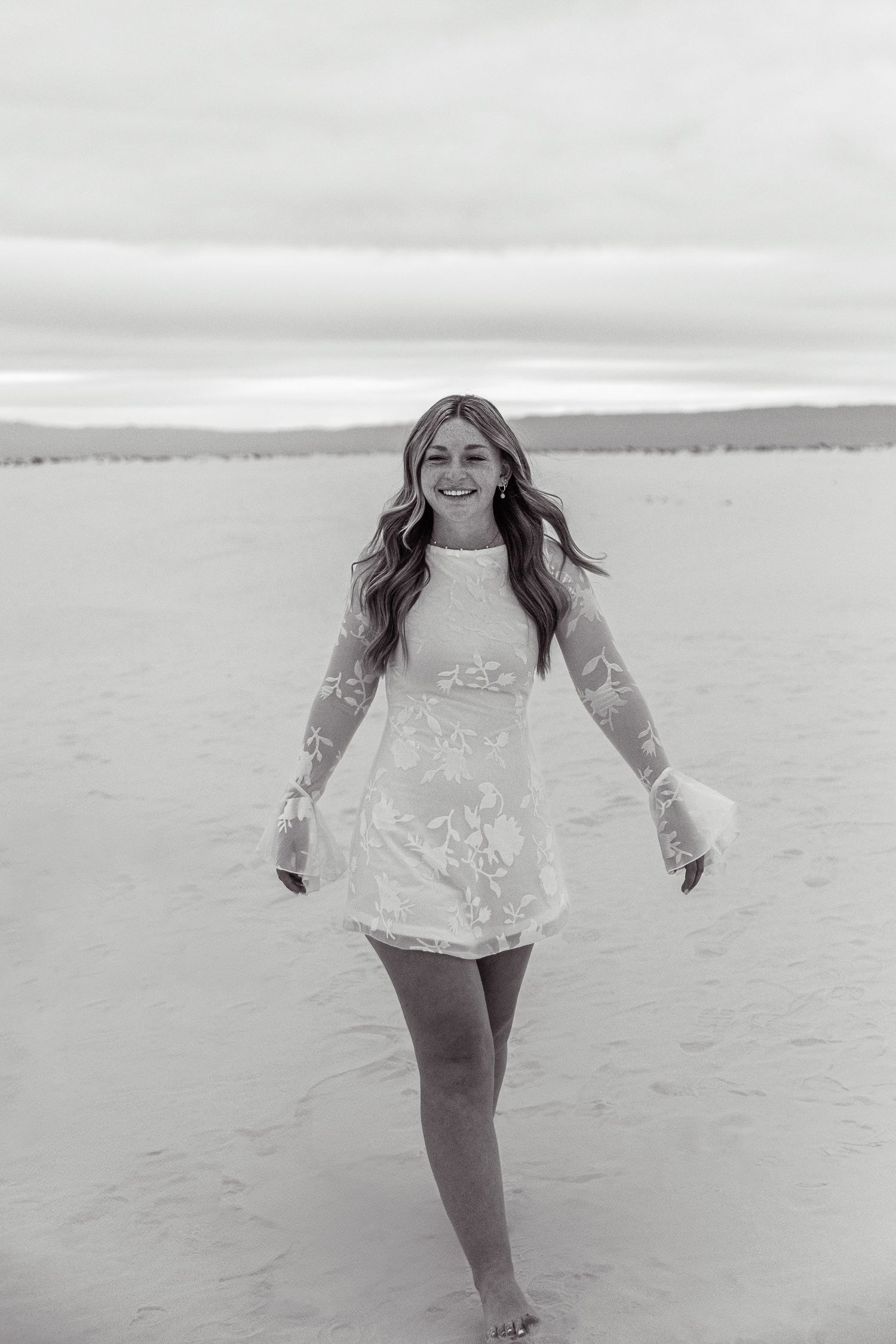 Black and white photo of a smiling young woman in a white lace mini dress walking barefoot on sand.