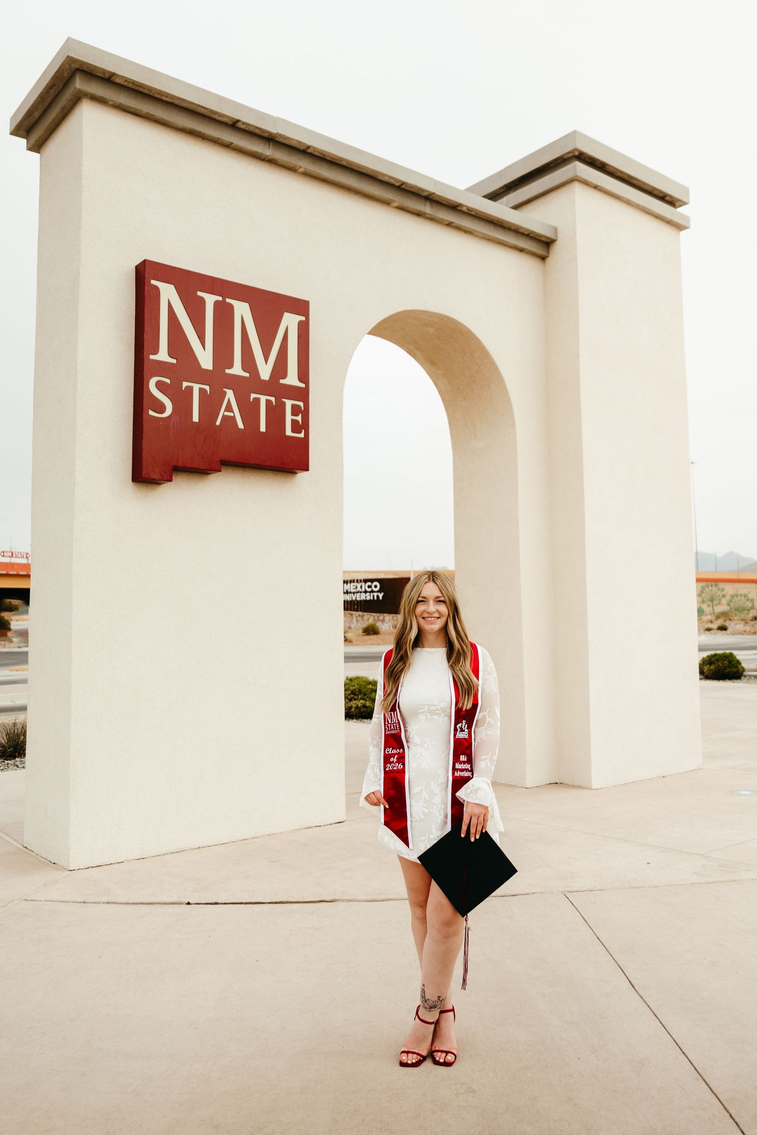 Graduating woman in white dress with red stole holding cap at NM State University arch.