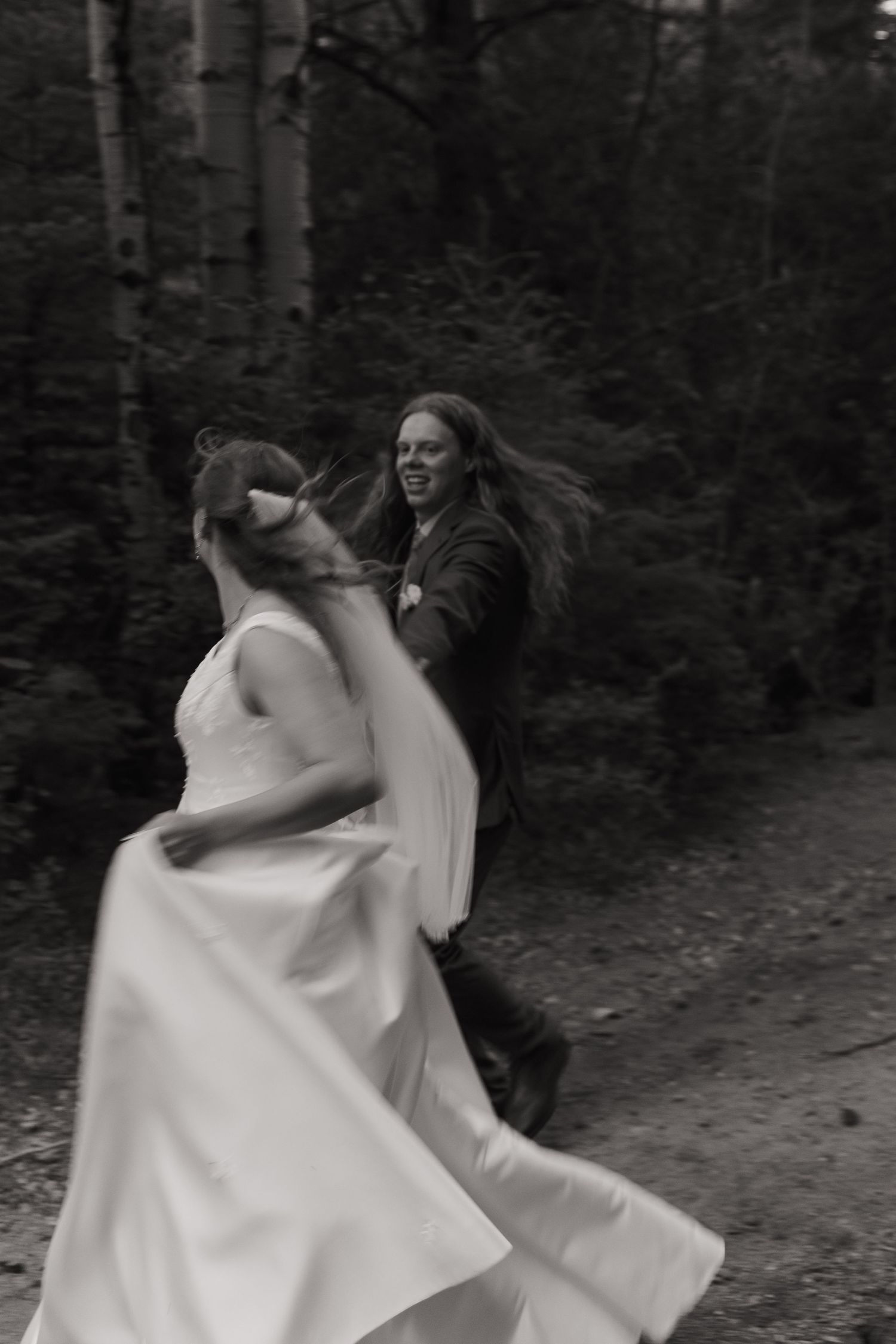 Black and white photo of a joyful bride and groom dancing together in a forest setting on their wedding day.