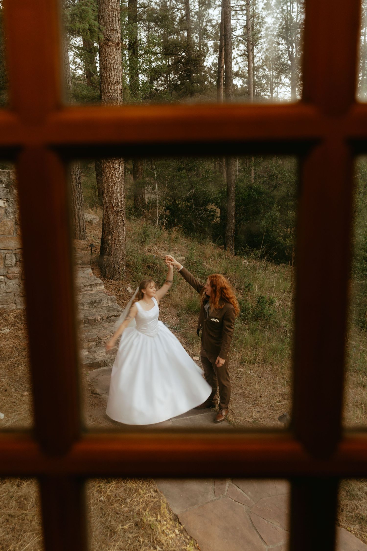 Bride and groom dancing in forest, viewed through rustic window frame, bride in white ball gown.
