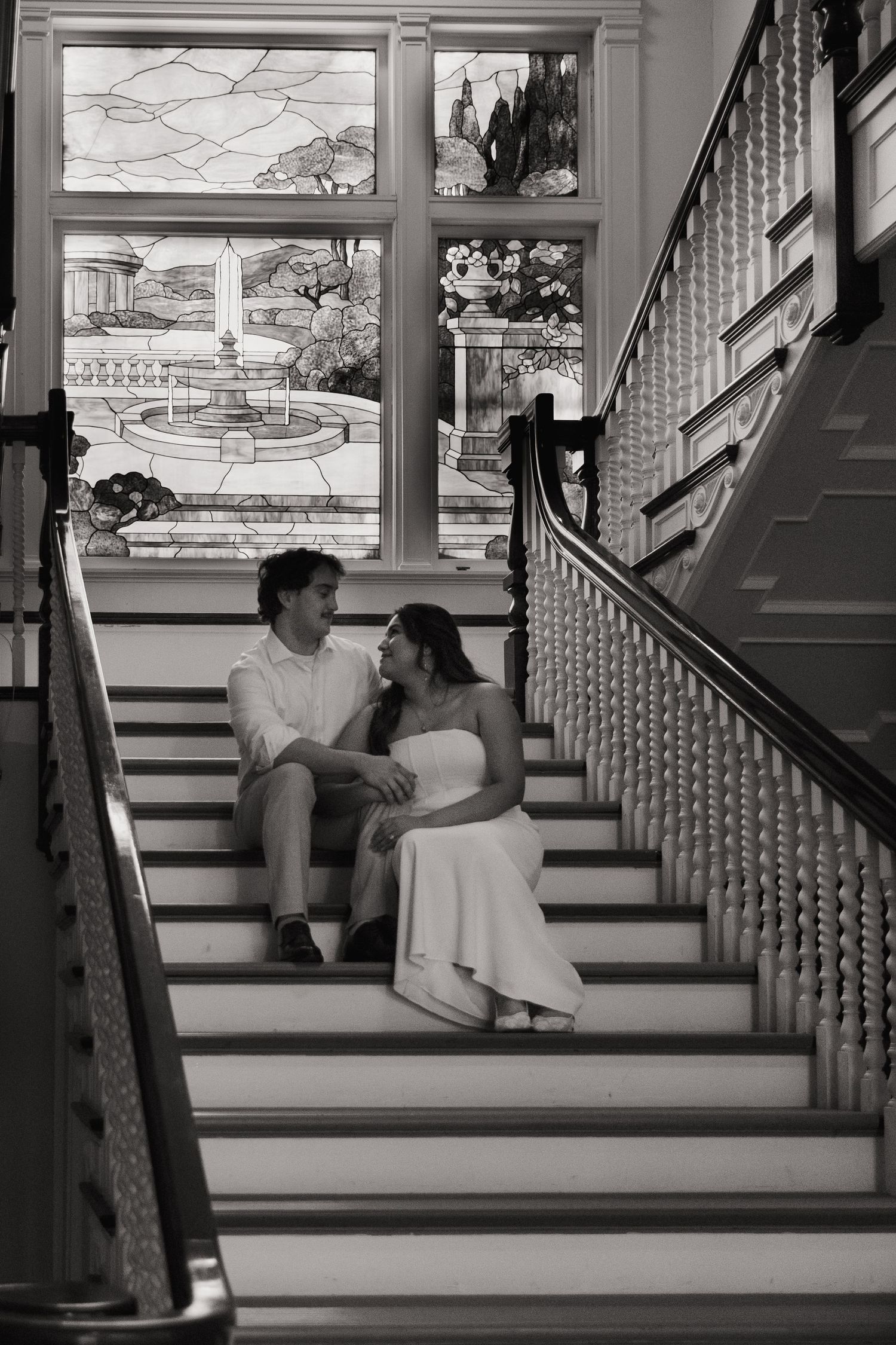 Couple sitting on elegant staircase steps in black and white photo with ornate stained glass window backdrop.