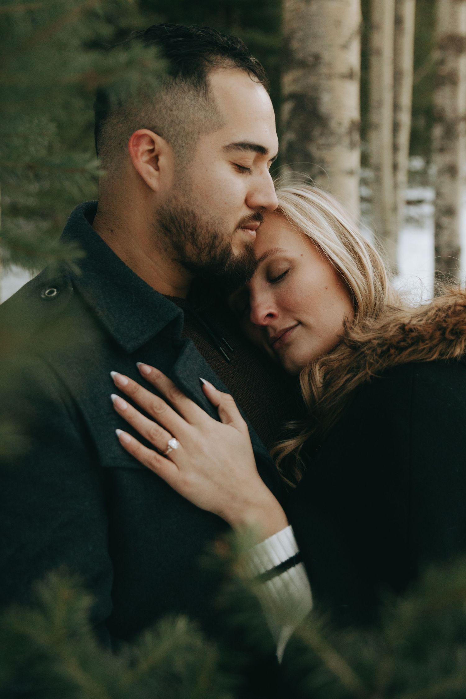Couple sharing a tender moment in a snowy forest, woman wearing engagement ring resting head on mans chest.