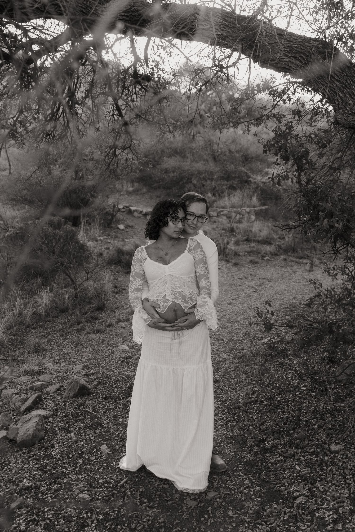 Black and white photo of a couple embracing outdoors, bride in white lace gown among desert trees.