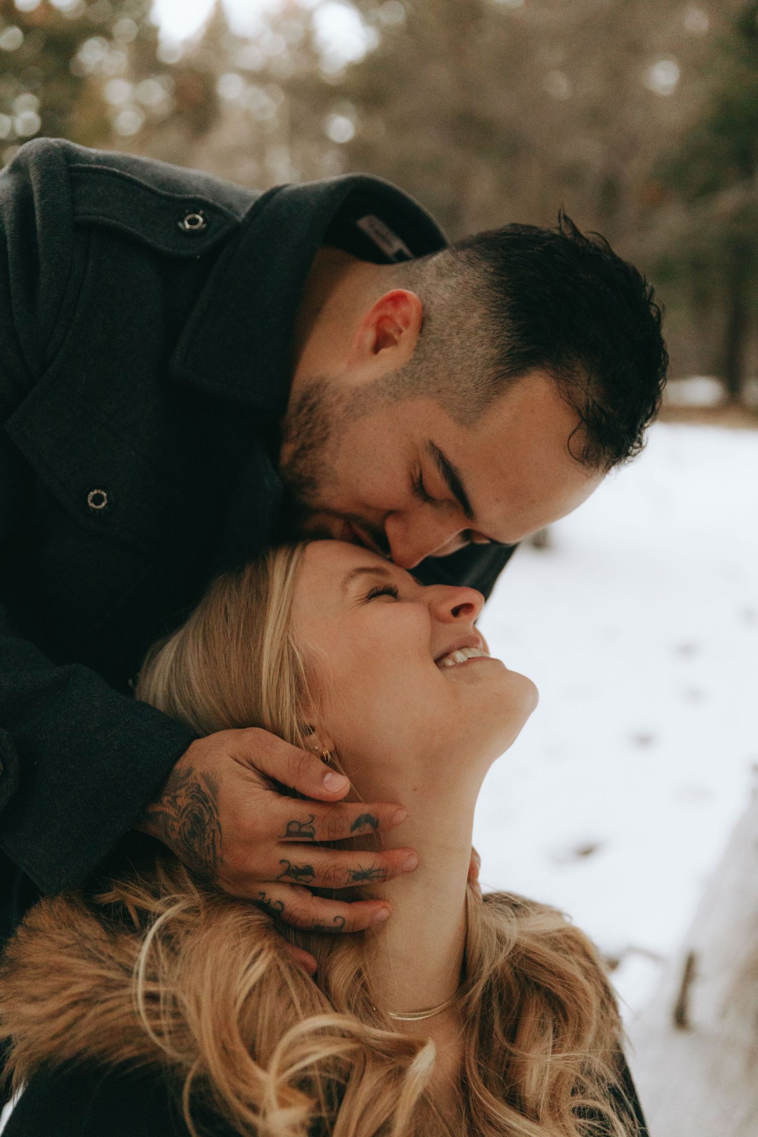 Couple sharing a romantic winter kiss outdoors, man with tattoos tenderly holding woman's face in snowy forest.