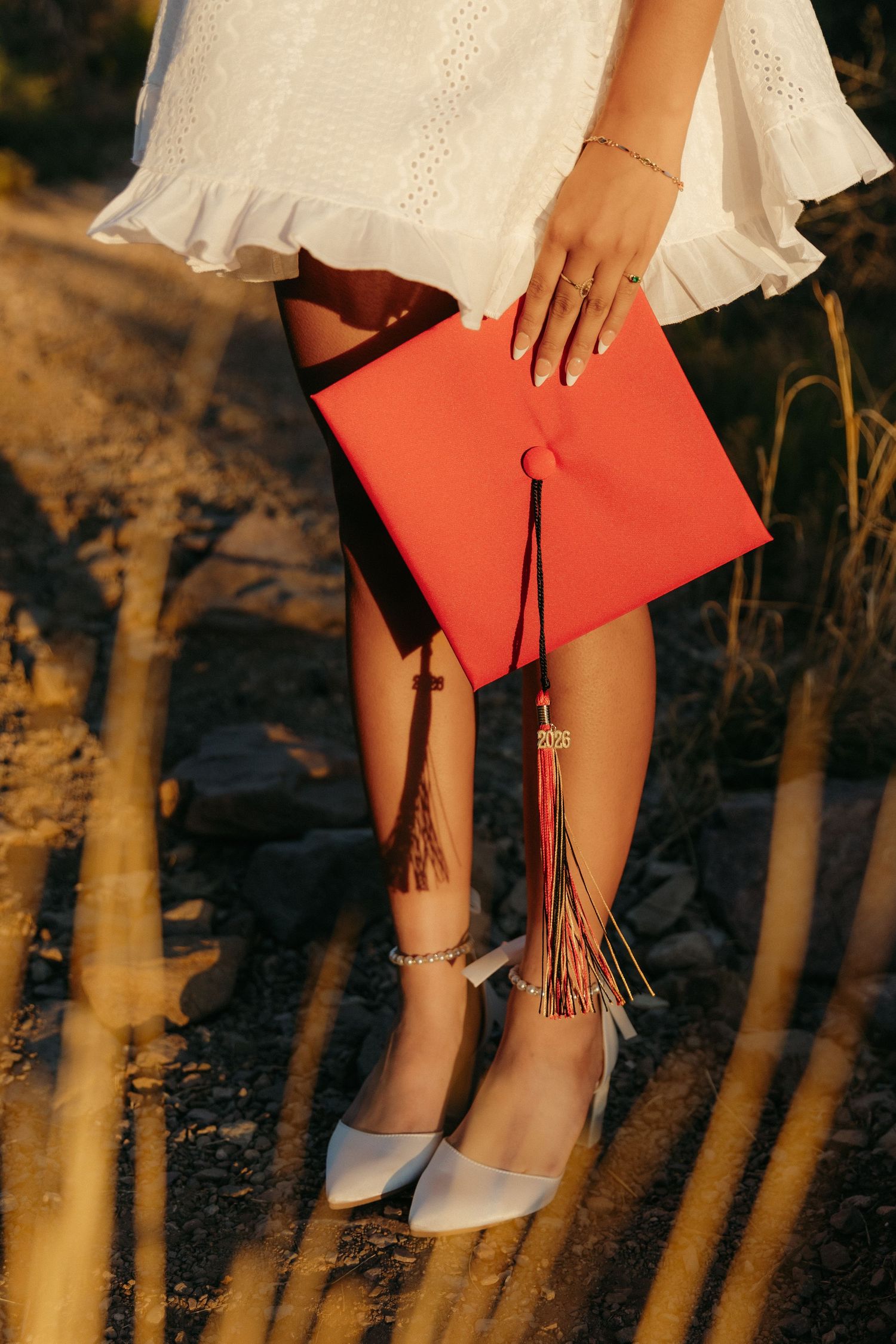 Close-up of graduation cap with red tassel held by woman in white dress and white heels outdoors.