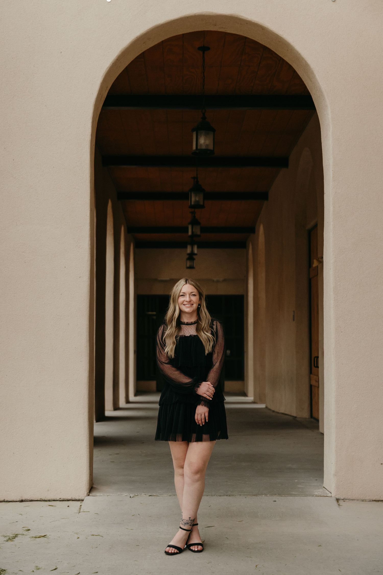 Young woman in black mini dress smiling under arched corridor colonnade.