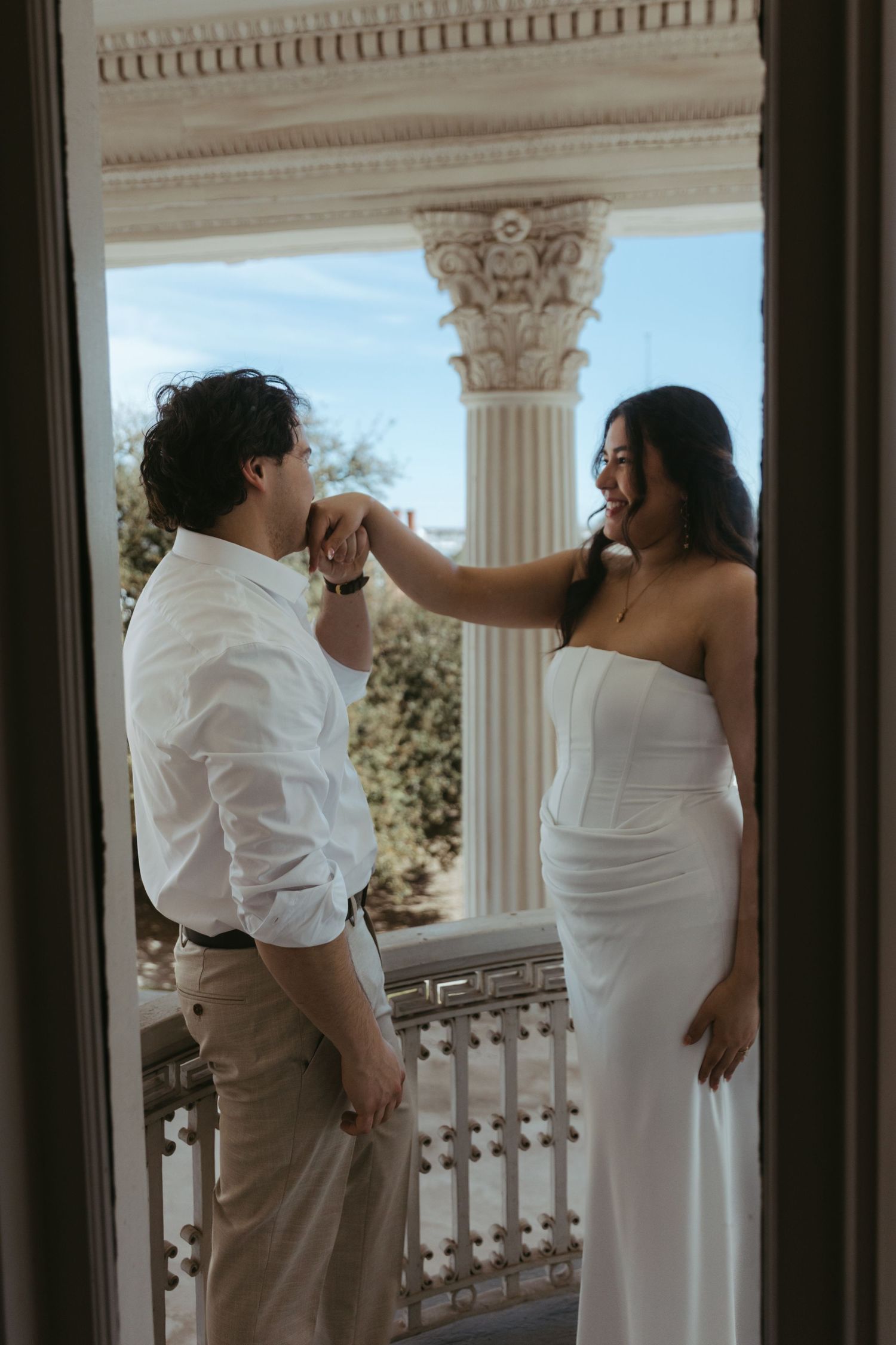 Groom kissing bride's hand on elegant columned balcony, bride in strapless white gown smiling.