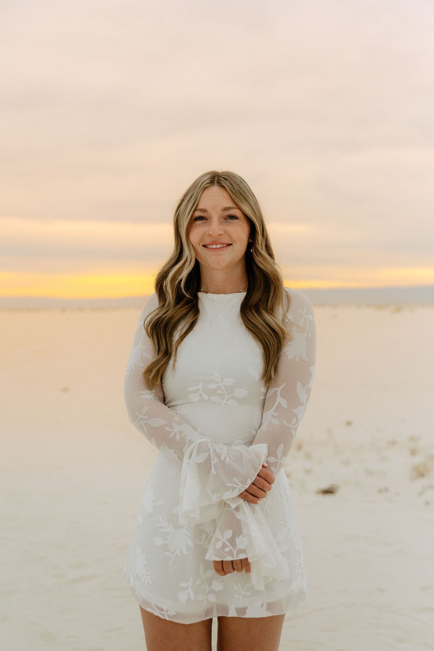 Young woman with long wavy blonde hair smiling in a white lace long-sleeve mini dress at a colorful sunset beach.