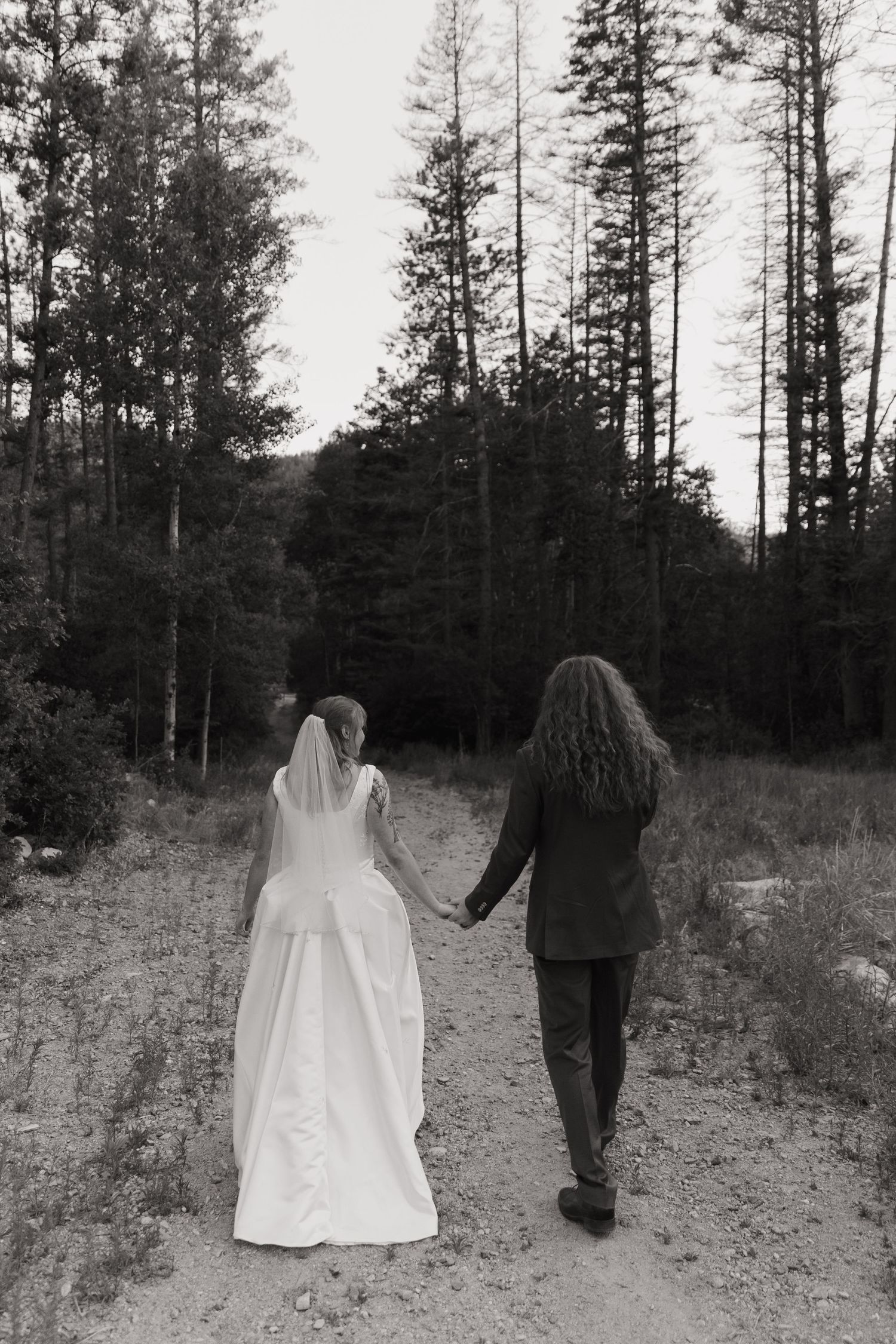 Black and white photo of bride and groom holding hands walking down a forest path on their wedding day.