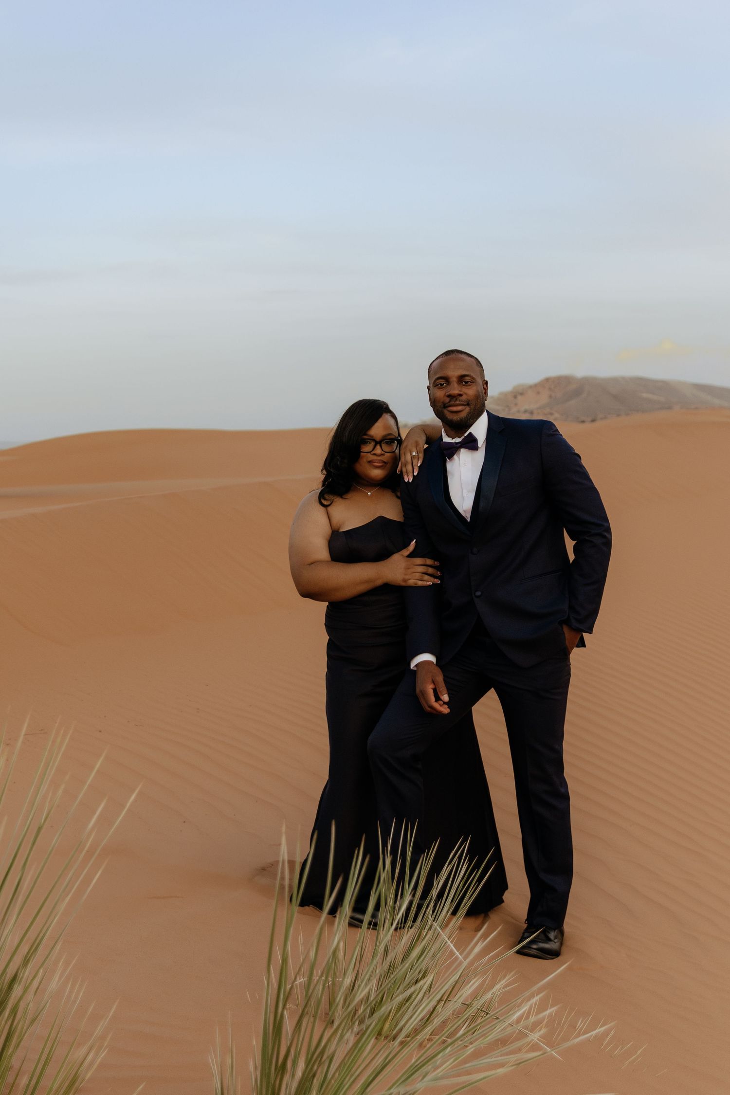 Couple in elegant black formalwear posing on sandy desert dunes during a romantic photoshoot.