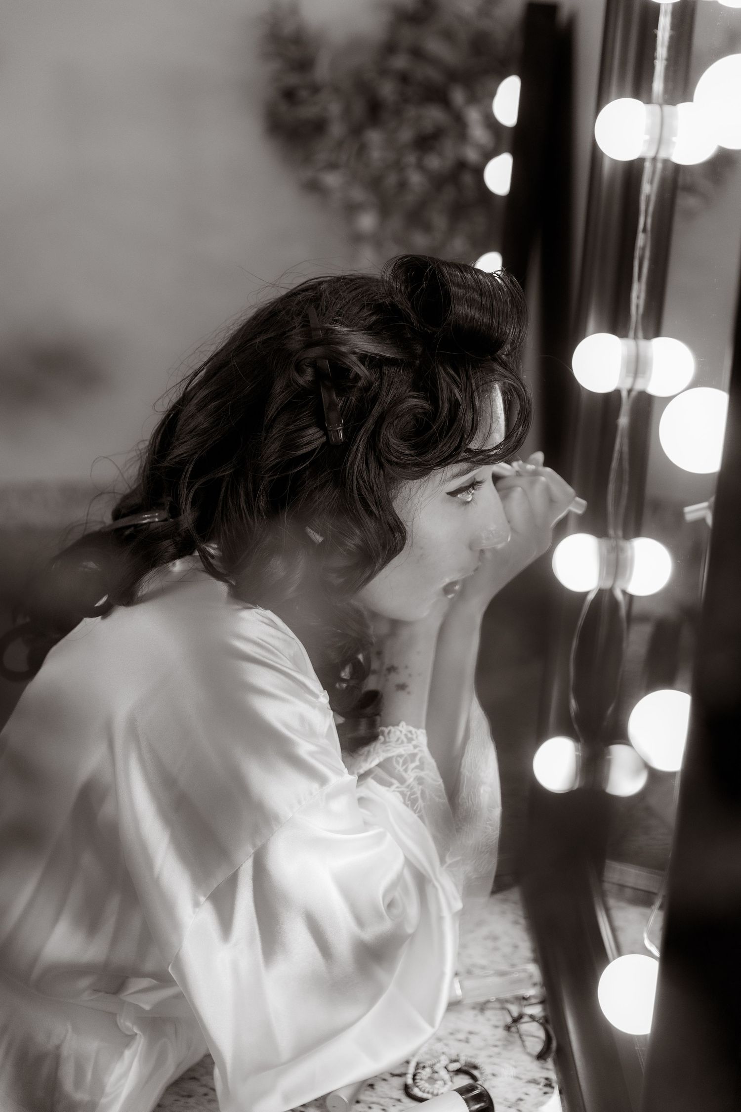 Black and white photo of a bride in silk robe applying makeup at a vanity mirror with glowing bulbs.