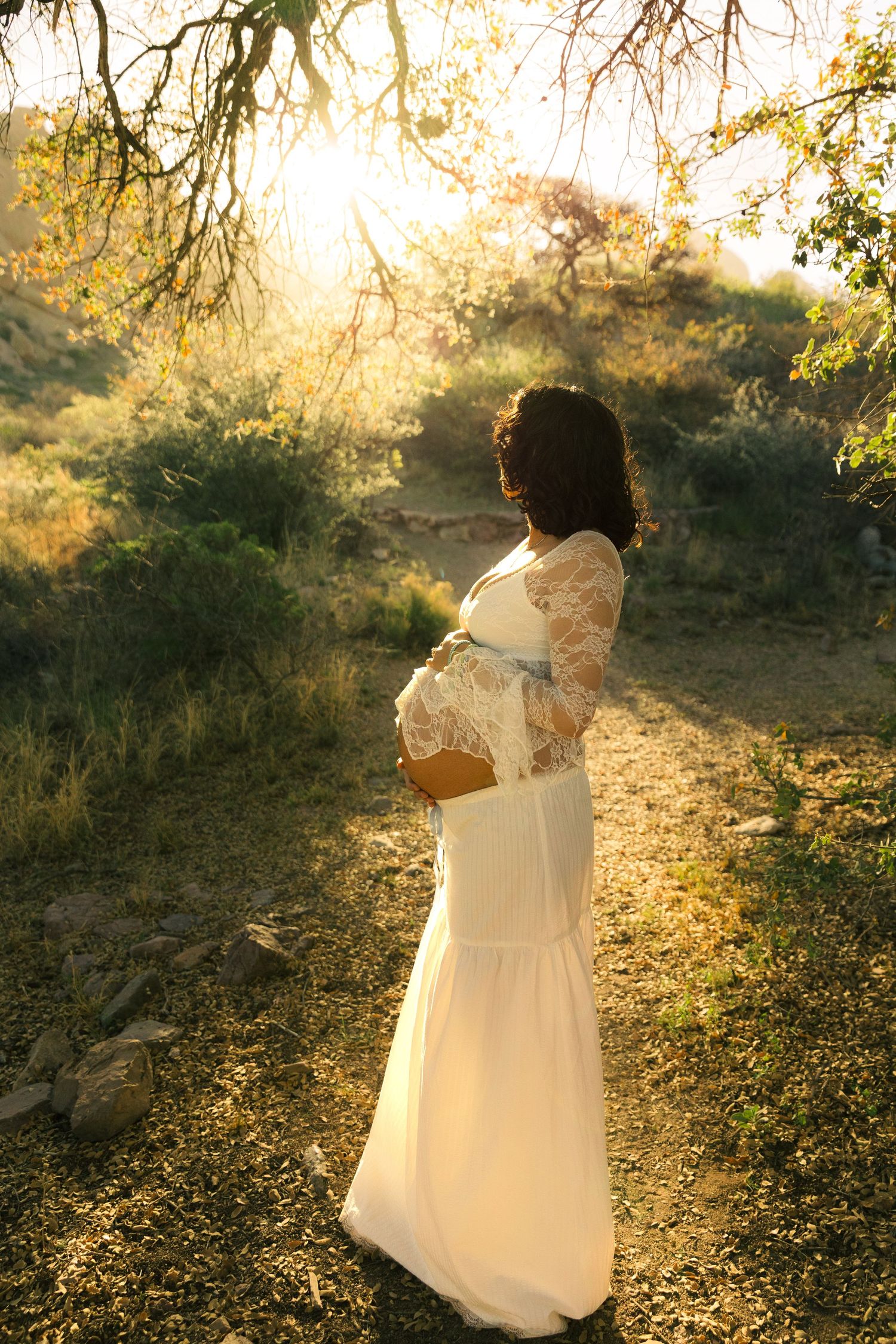 Pregnant woman in white lace gown cradling belly outdoors in golden sunlight among lush greenery.