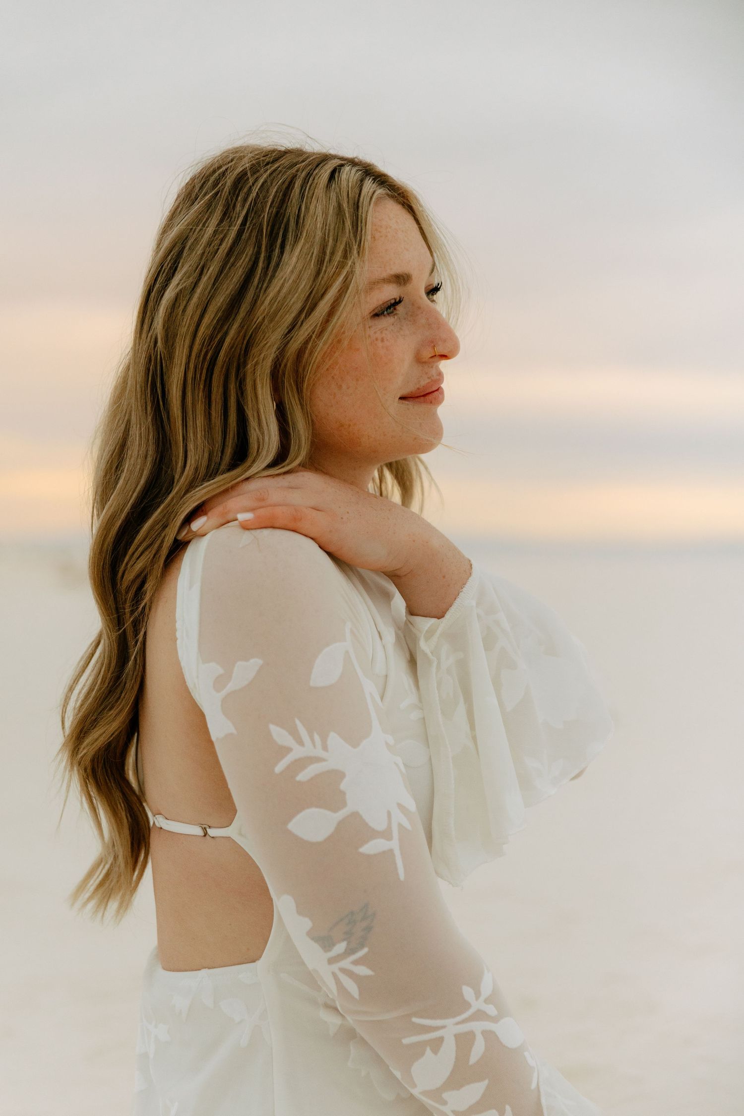 Close-up side profile of woman in white floral lace backless dress at sunset beach.
