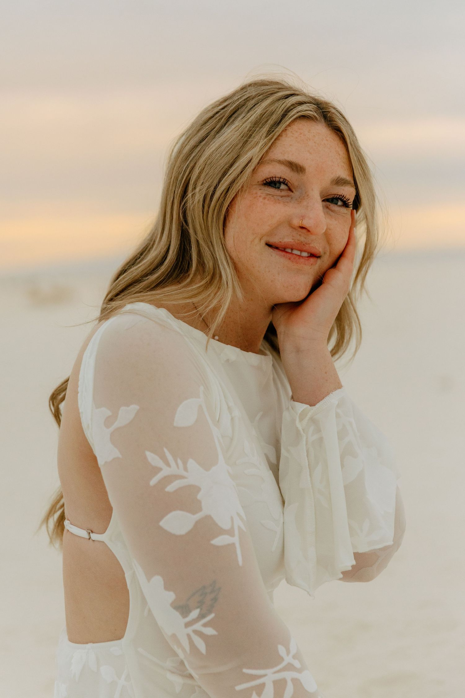 Freckled blonde woman in white lace dress rests cheek on hand smiling softly on sandy beach.