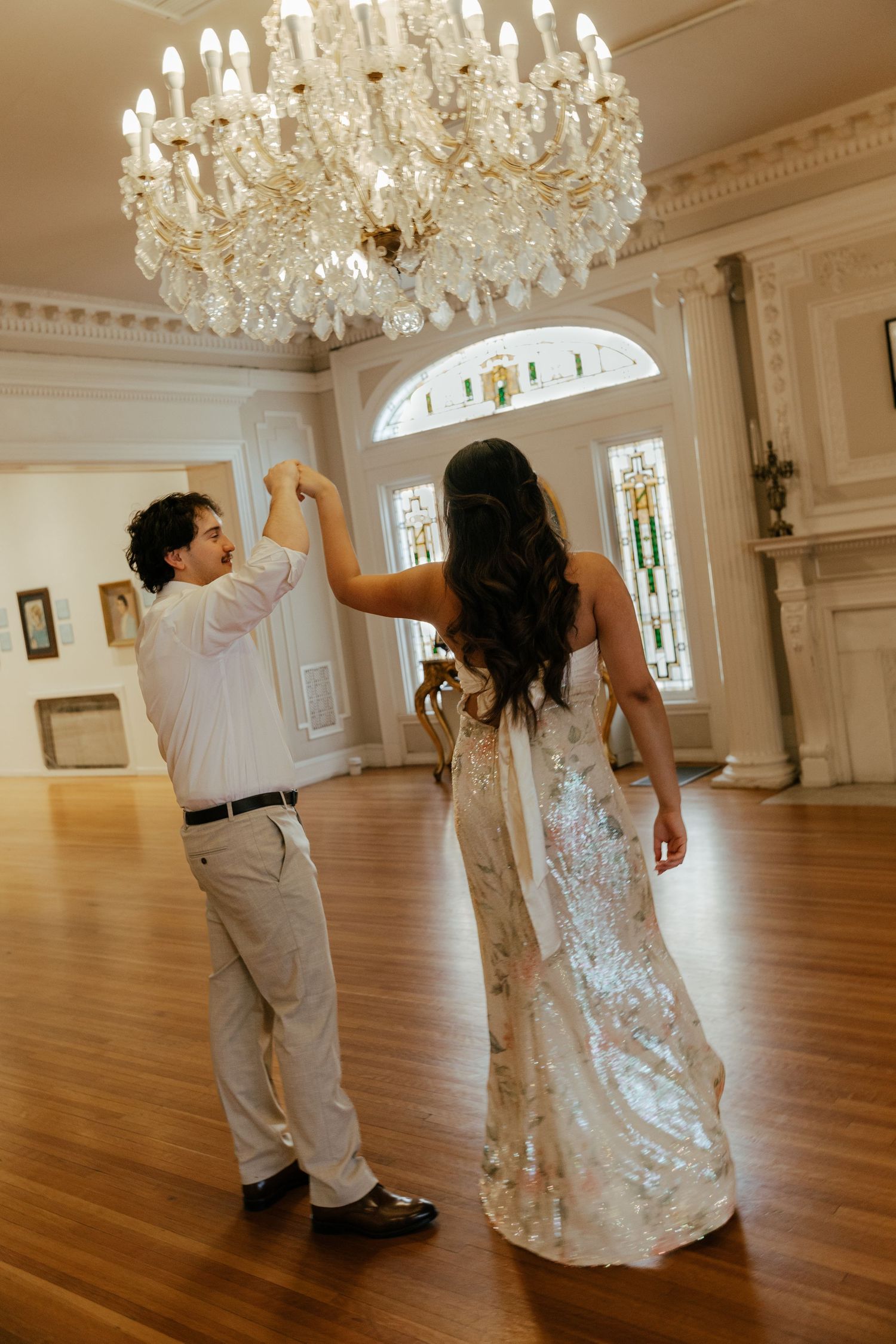 Bride and groom share a first dance under a crystal chandelier in a grand ballroom with stained glass windows.