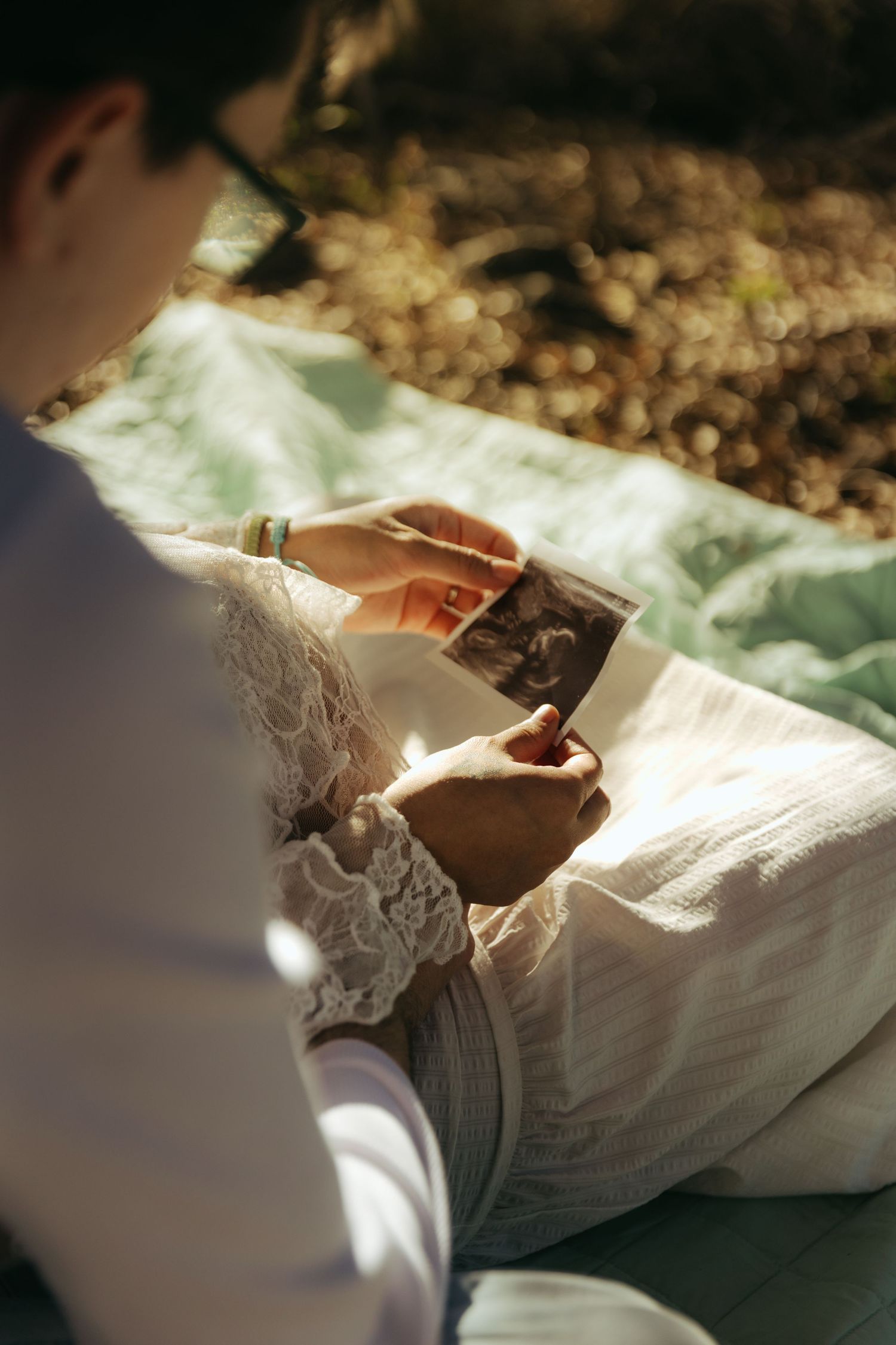 Pregnant woman in white lace dress sitting outdoors on blanket, looking at ultrasound photo in sunlight.