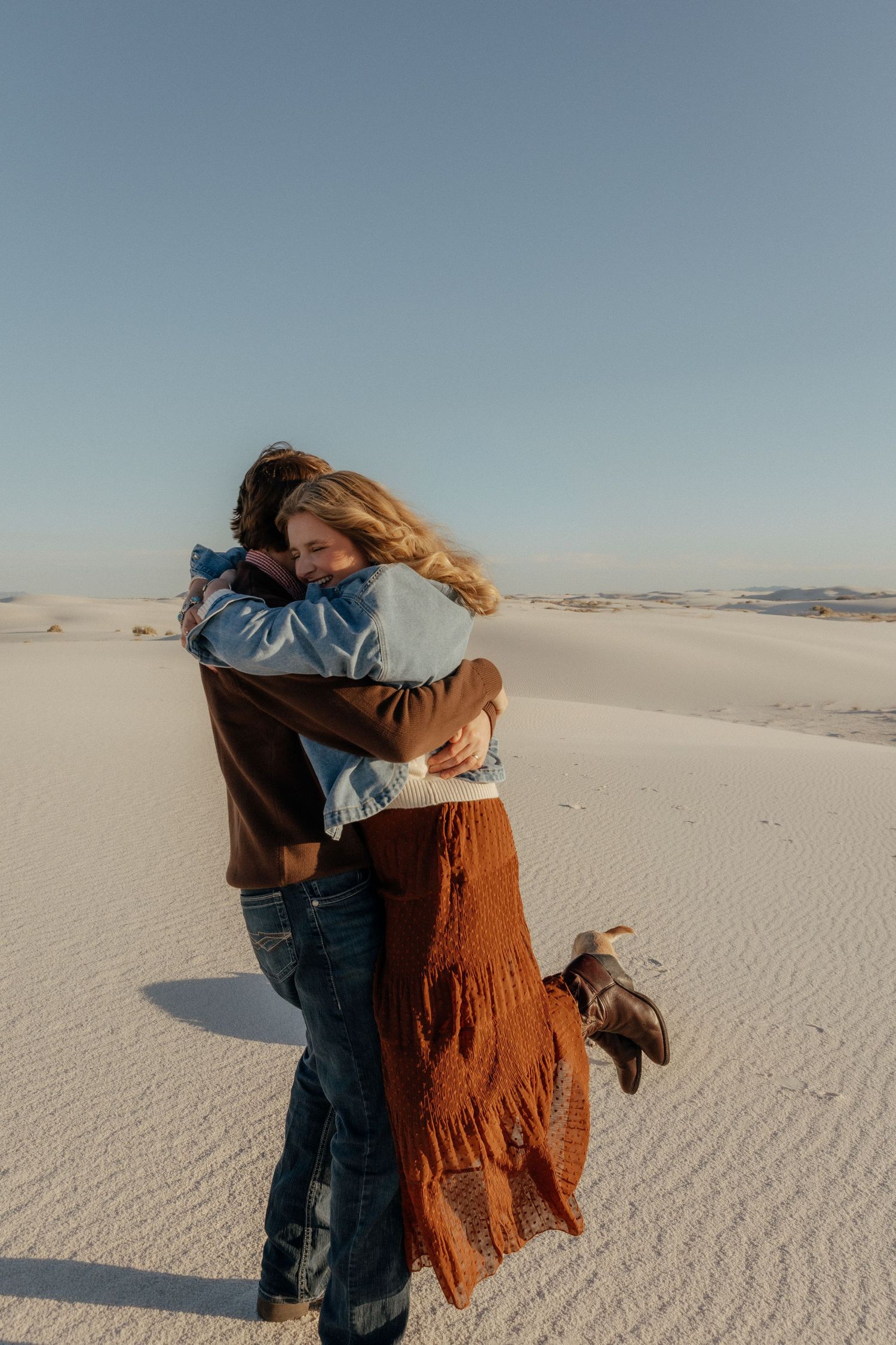Two people sharing a romantic hug on expansive white sand dunes at golden hour.