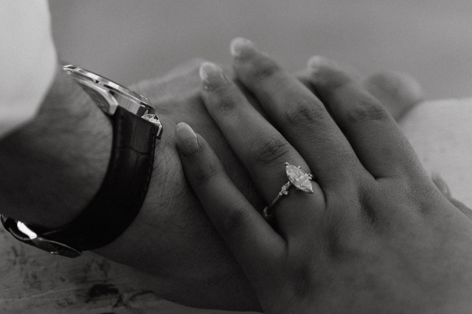 Black and white photo of a couple holding hands, woman wearing a diamond engagement ring, man wearing a leather watch.