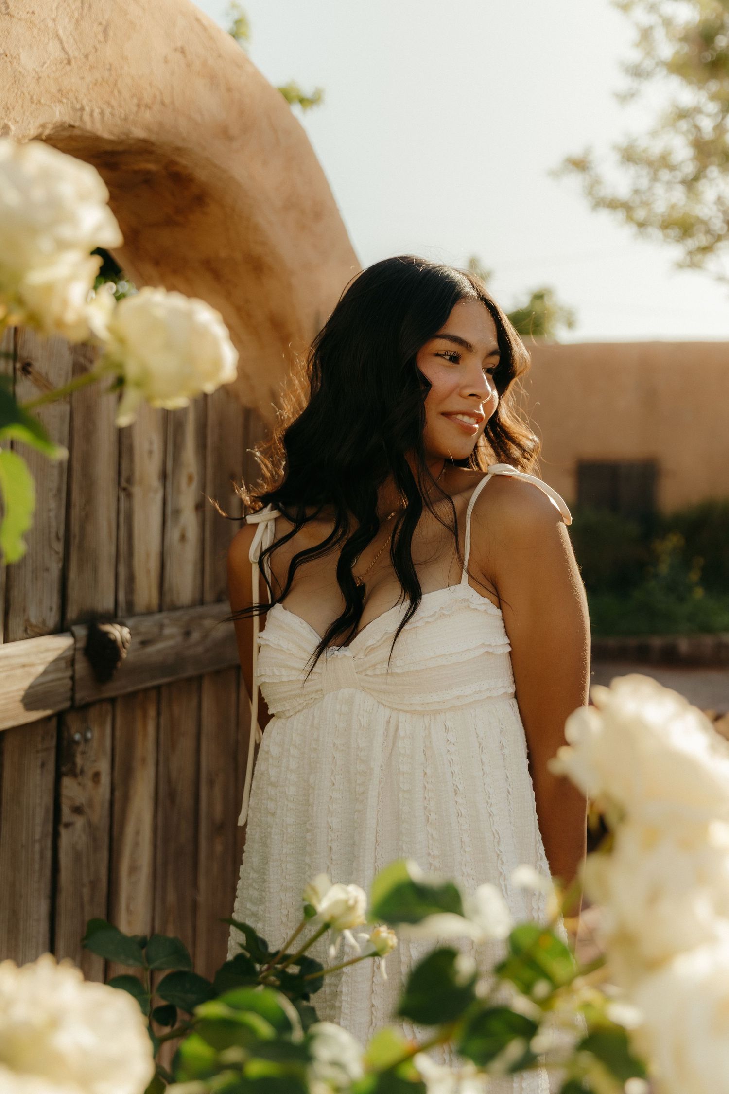 A young woman in a white sundress stands by a rustic wooden door surrounded by white roses in warm golden light.