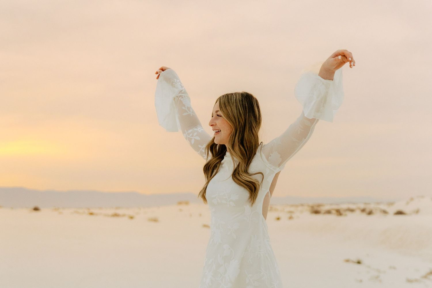 Joyful young woman in a white lace dress raising her arms up against a soft pastel sunset sky on a sandy beach.