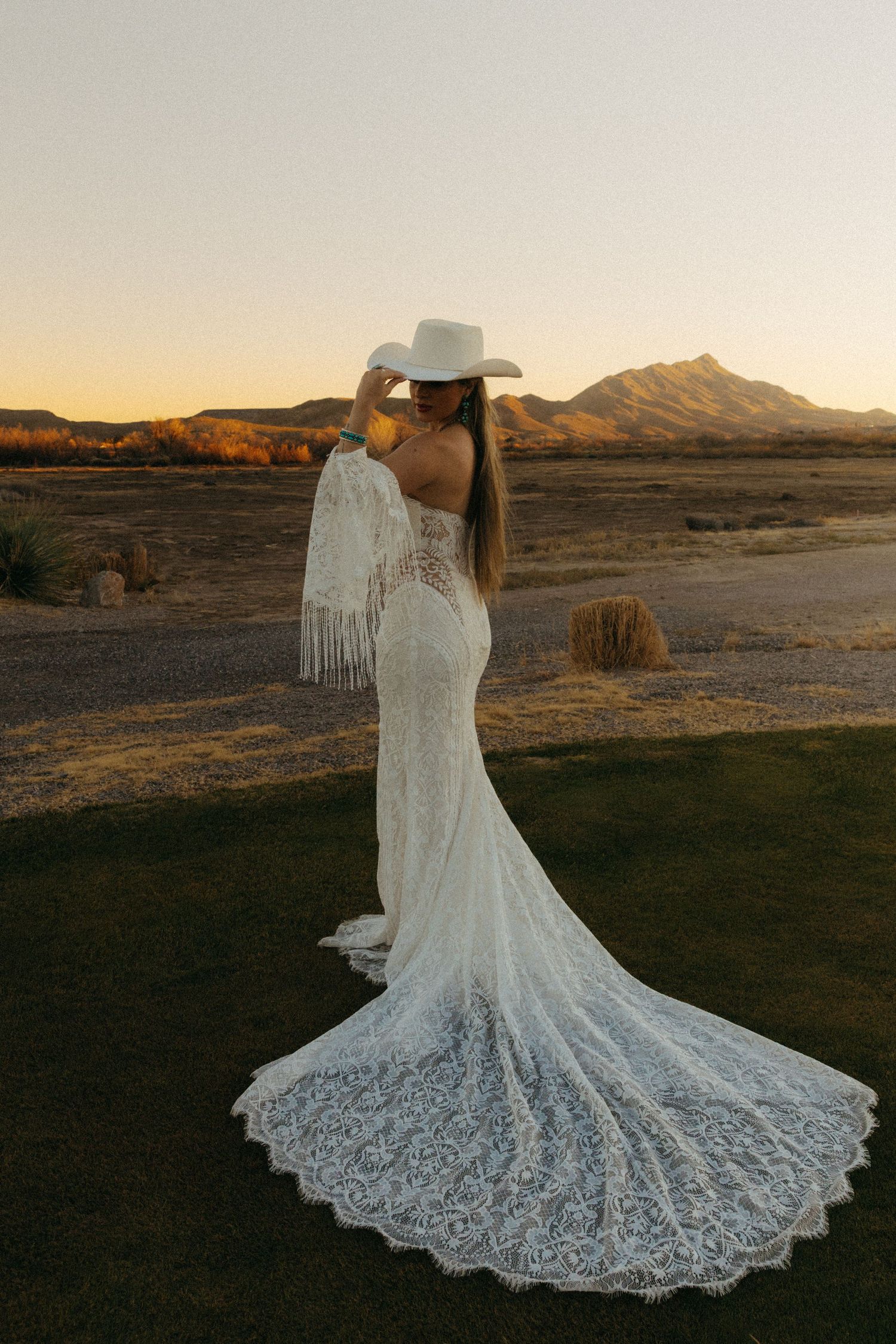 Bride in backless lace mermaid gown and white cowboy hat poses at desert sunset with mountain backdrop.