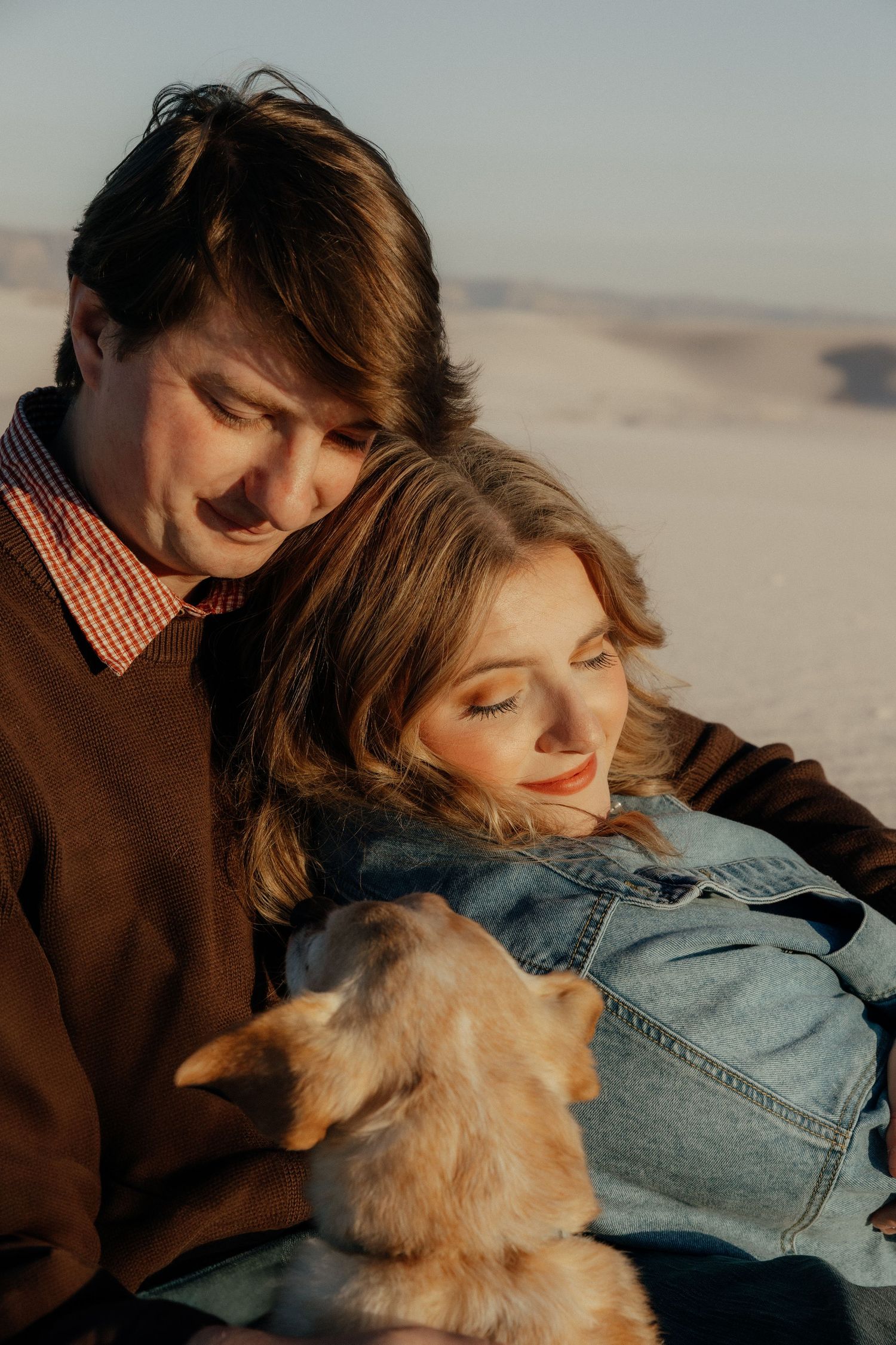 Couple sitting together on white sand dunes at golden hour, woman resting head on mans shoulder with a small dog.
