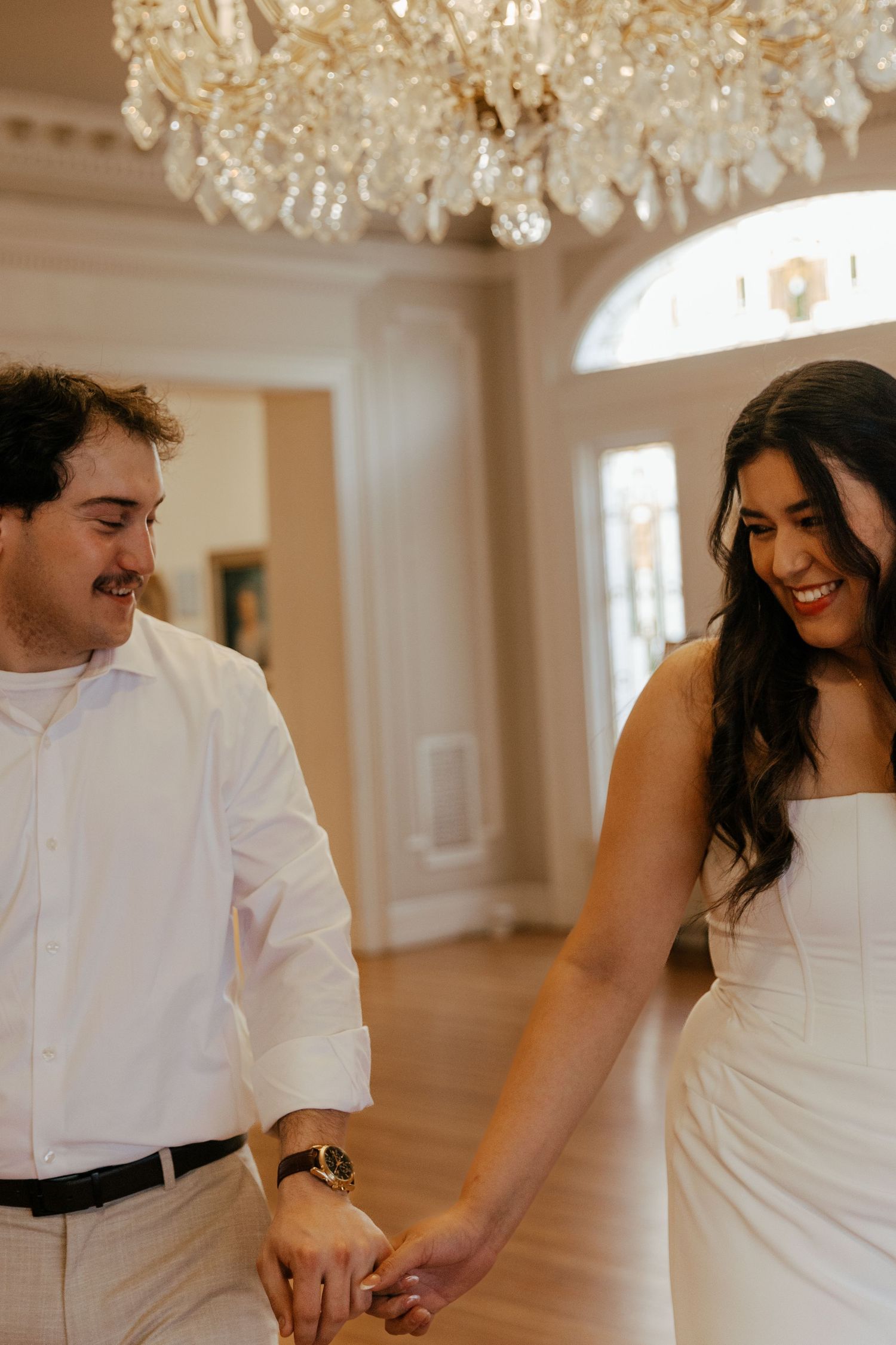 Smiling couple in white attire holding hands under a crystal chandelier in an elegant venue.