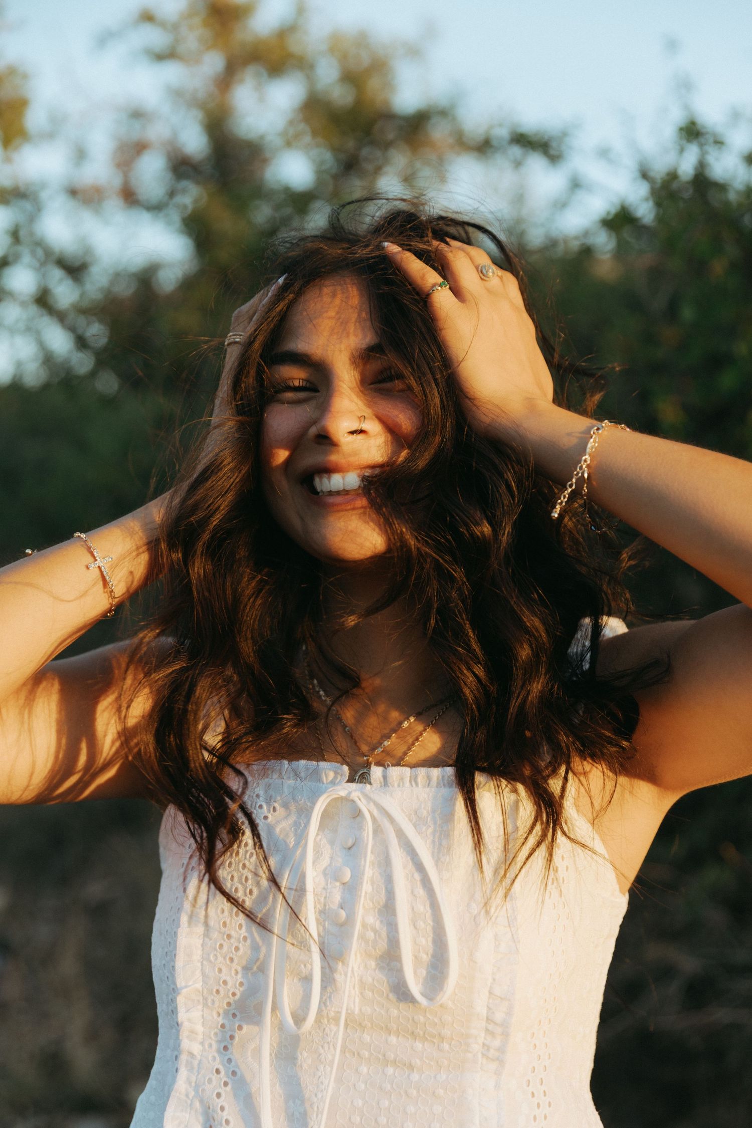 Smiling young woman in white sundress with wavy brown hair, hands in hair, golden hour sunlight outdoors.