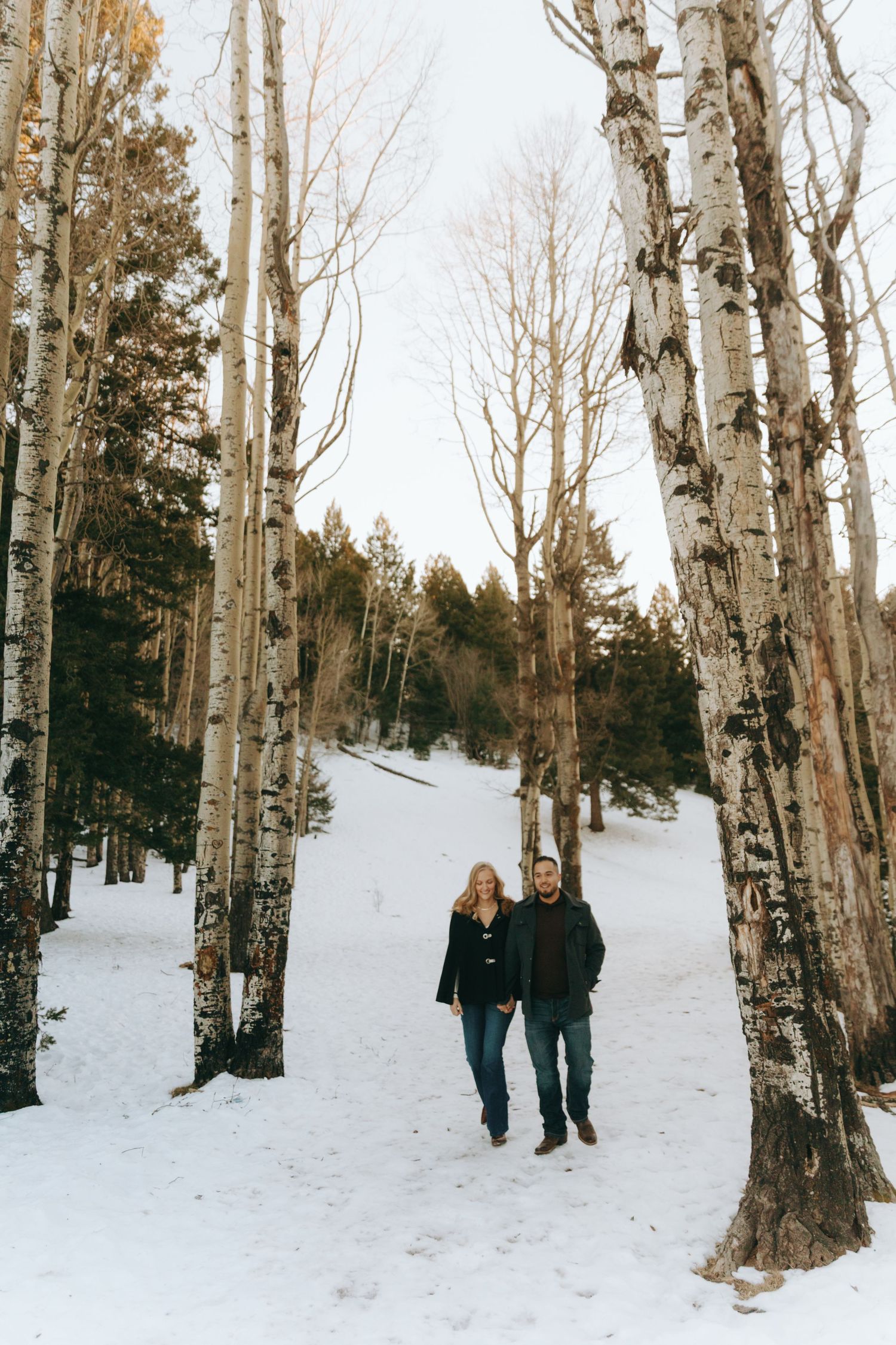 Couple walking hand in hand through snowy birch tree forest in winter.