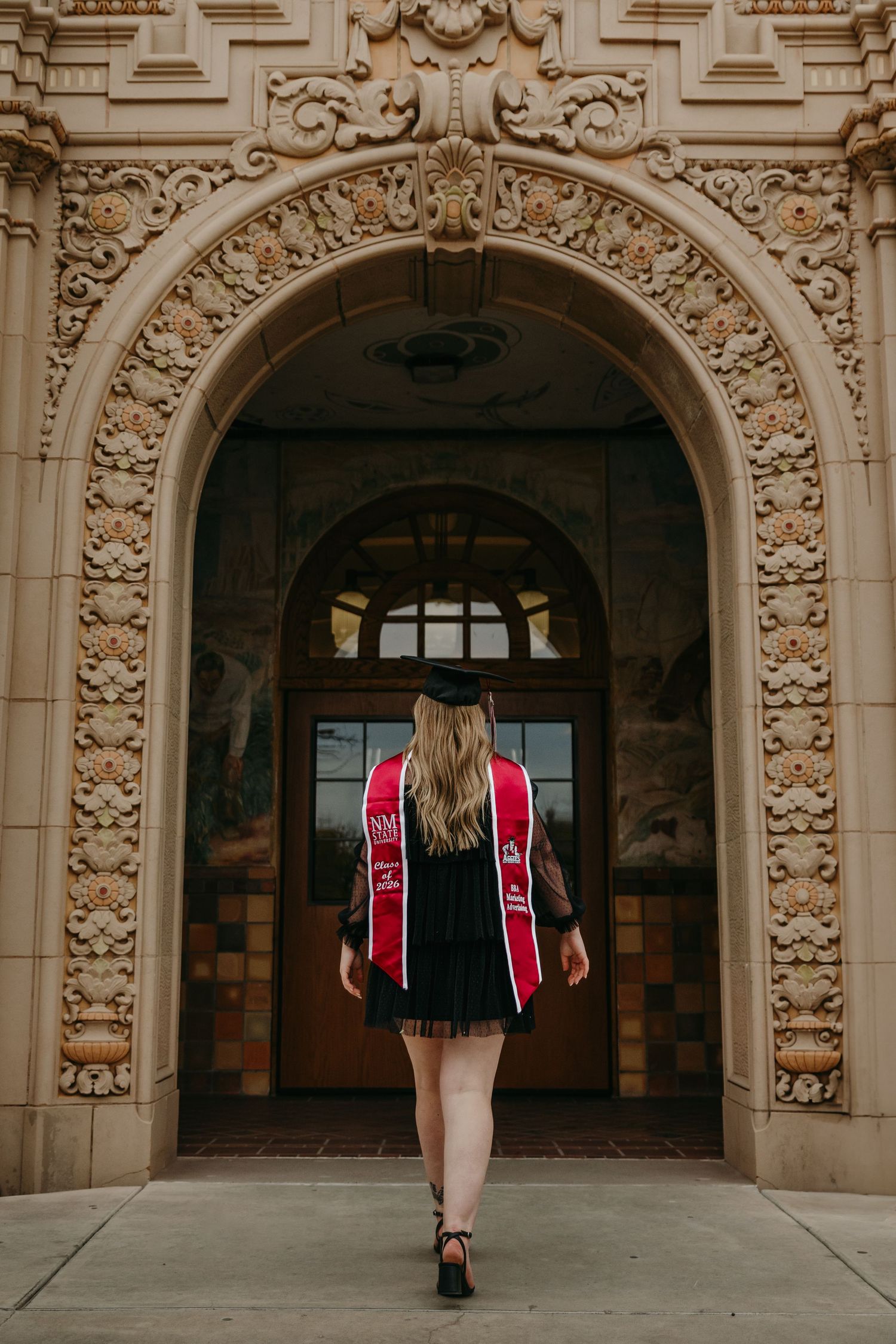 Graduate with red stole and cap walking through elaborate carved stone archway.