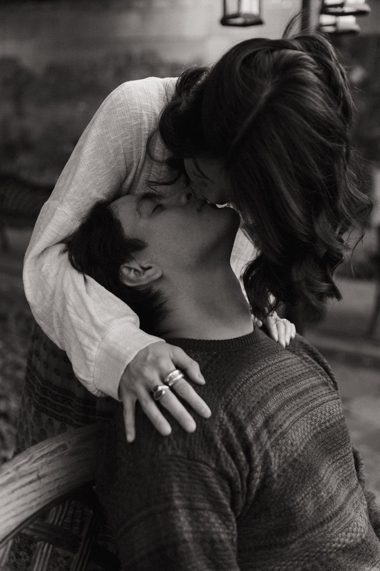 Black and white photo of a woman in a loose knit top leaning down to kiss a seated man in a textured sweater, her hand resting on his chest.
