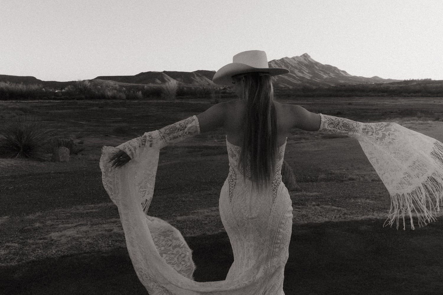 Black and white photo of bride in lace wedding dress and cowboy hat with arms outstretched in desert.
