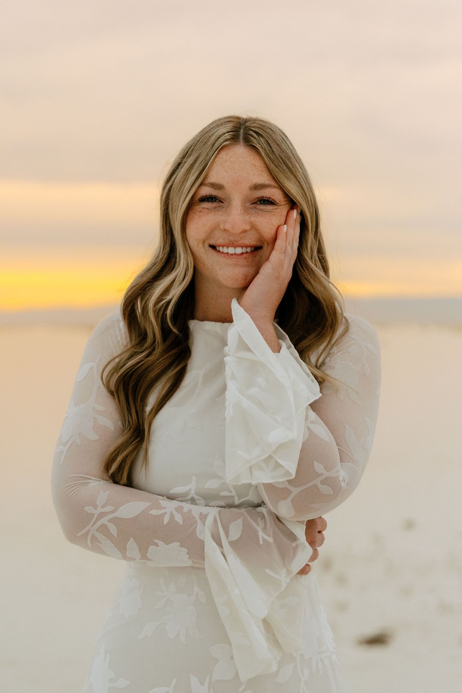 Smiling young woman in a white lace dress resting her hand on her cheek during a golden sunset beach photoshoot.