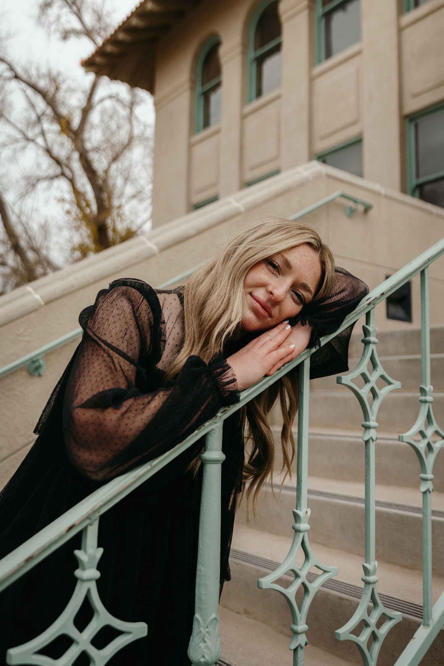 Blonde woman in black sheer dress leaning on mint green ornate stair railing.