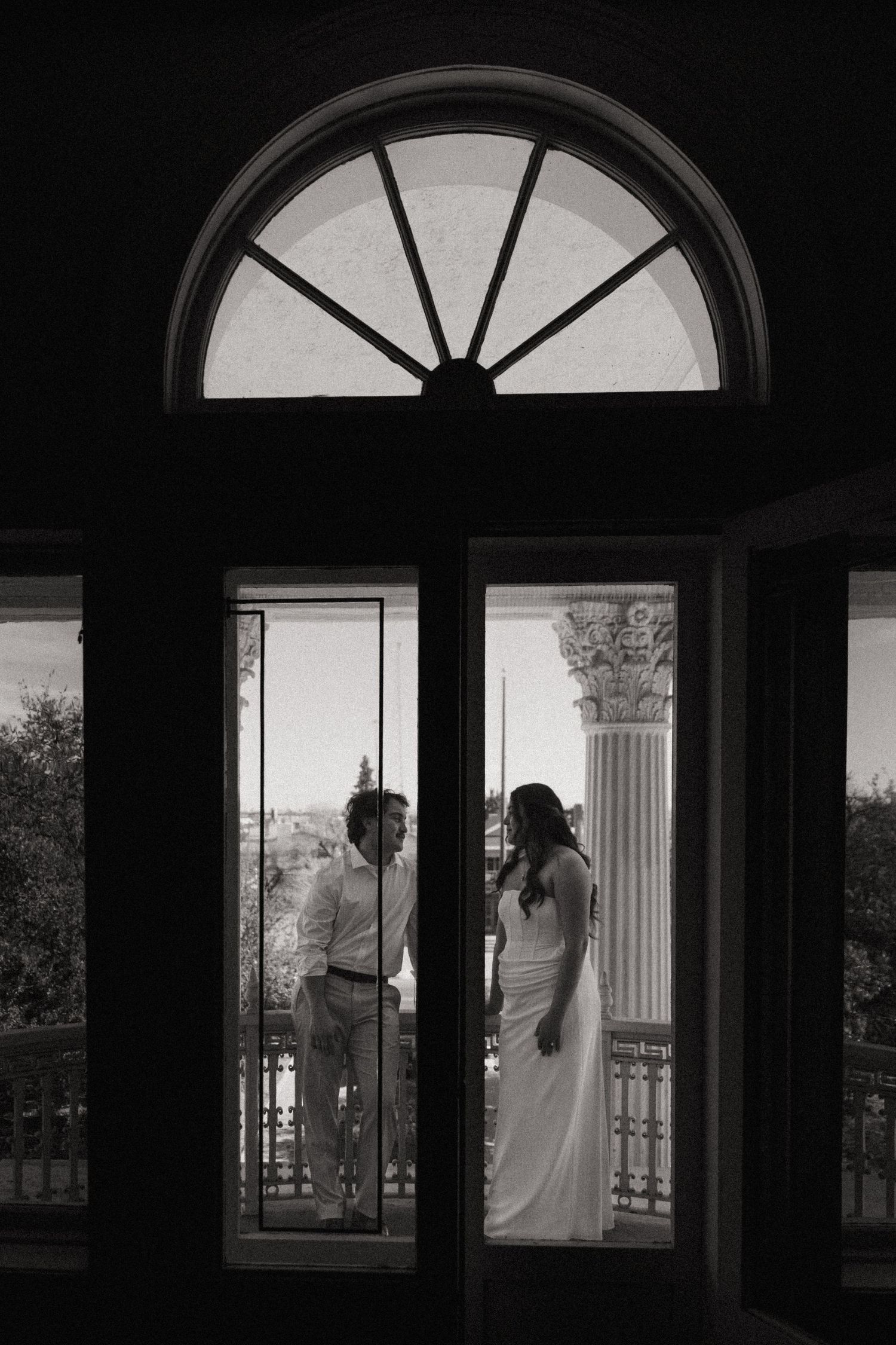 Black and white photo of bride and groom standing on a balcony framed by elegant arched French doors.