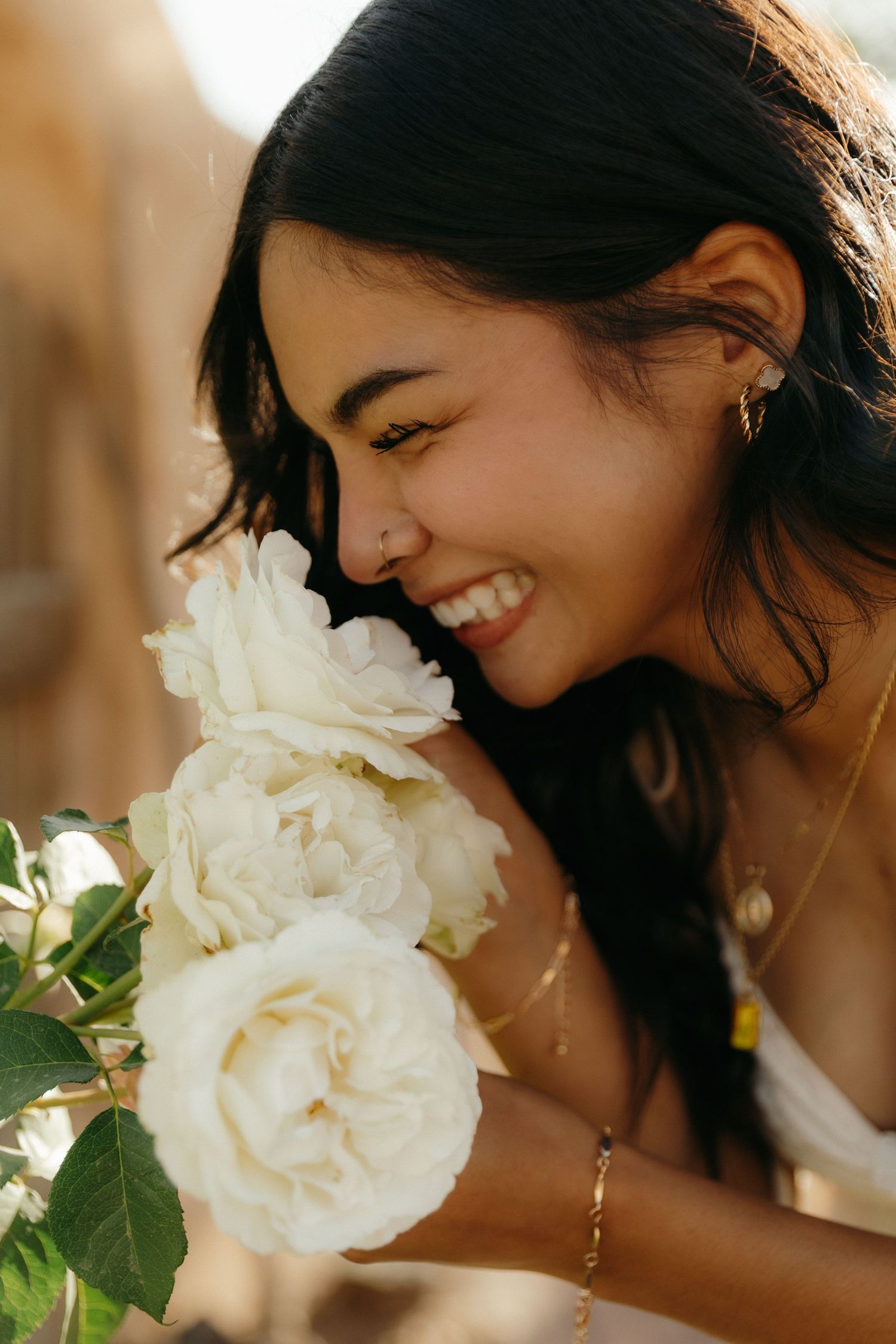 Smiling young woman with dark hair smelling white roses, wearing gold jewelry in warm sunlight.