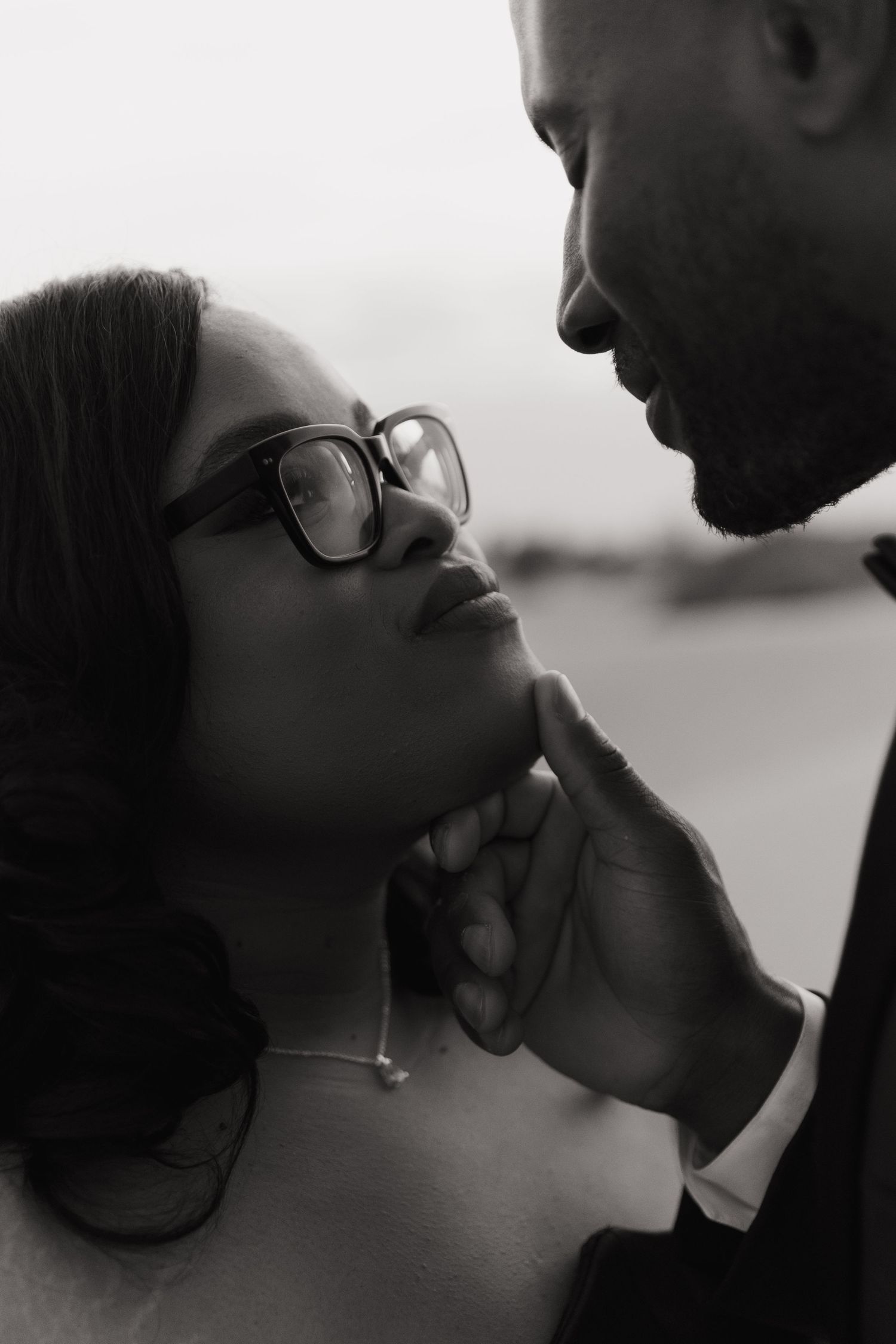 Black and white romantic couple portrait, man gently lifting woman's chin, woman wearing glasses, intimate wedding moment.