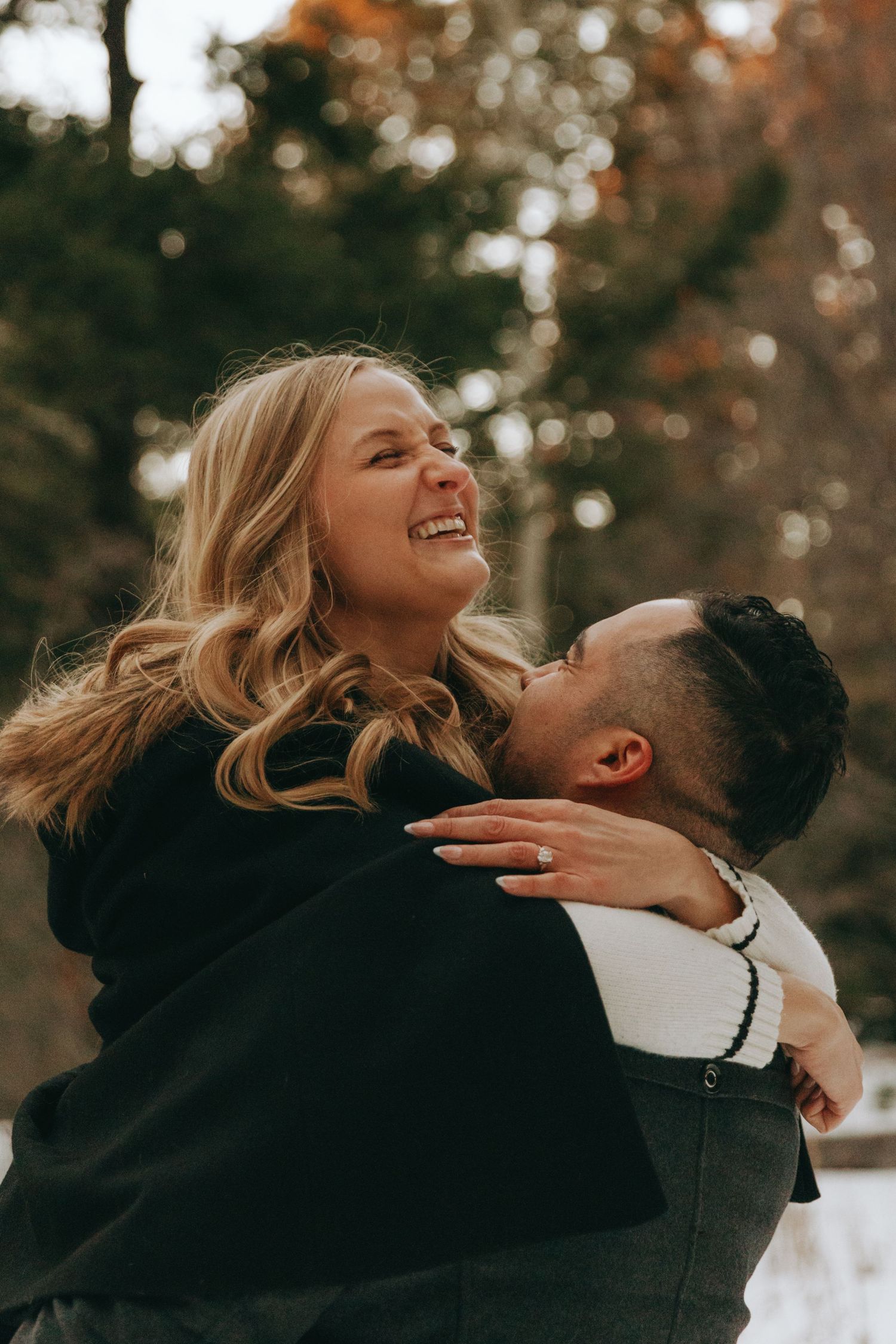 A joyful couple embraces outdoors in autumn, woman laughing as man lifts her, showcasing an engagement ring.