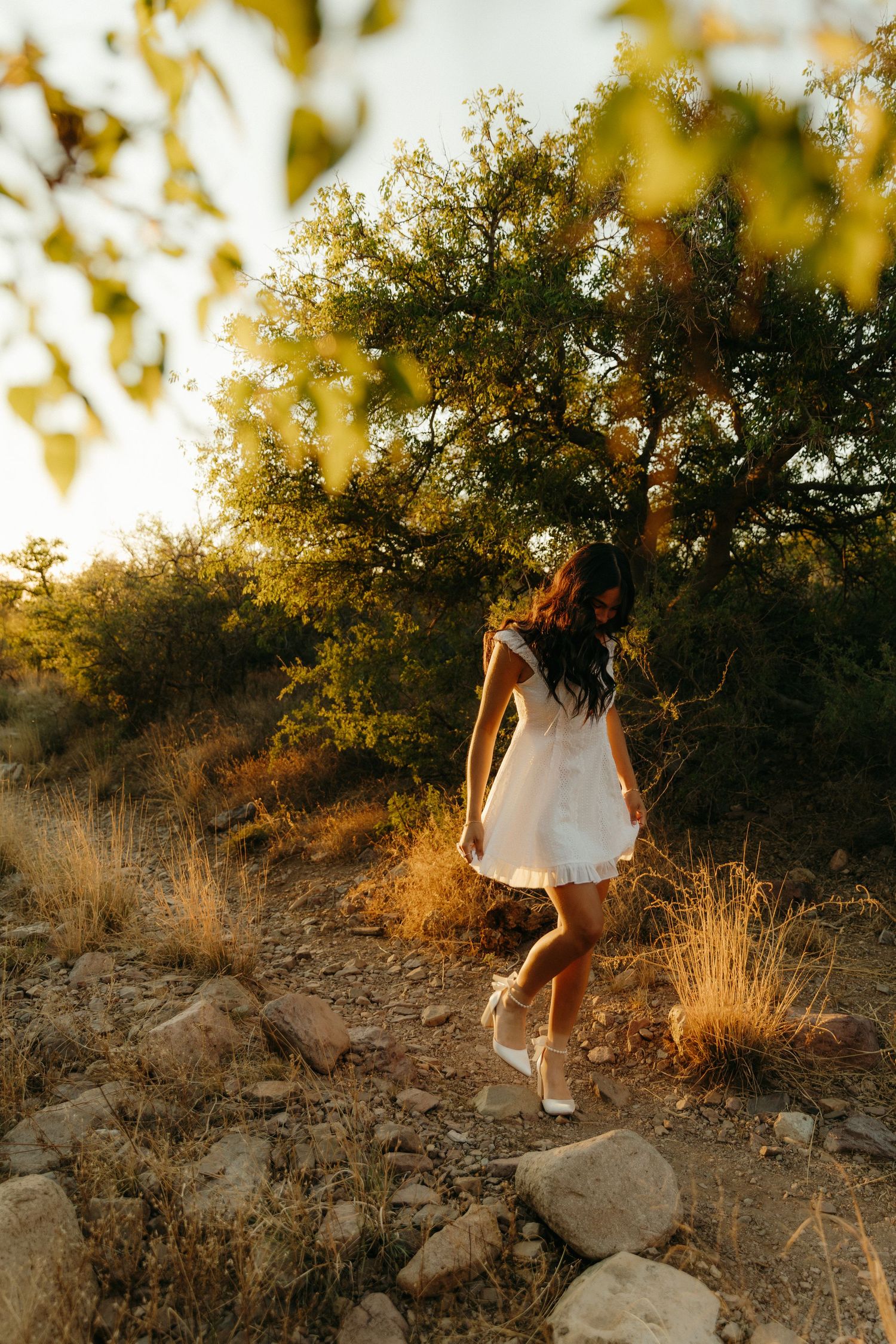 Young woman in white mini dress walking on rocky trail surrounded by trees in warm golden sunset light.