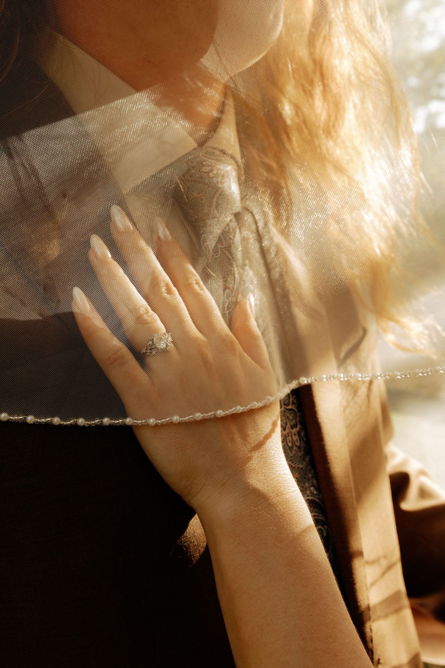 Woman's hand adorned with a diamond ring and delicate bracelet, draped in sheer fabric with warm golden light.
