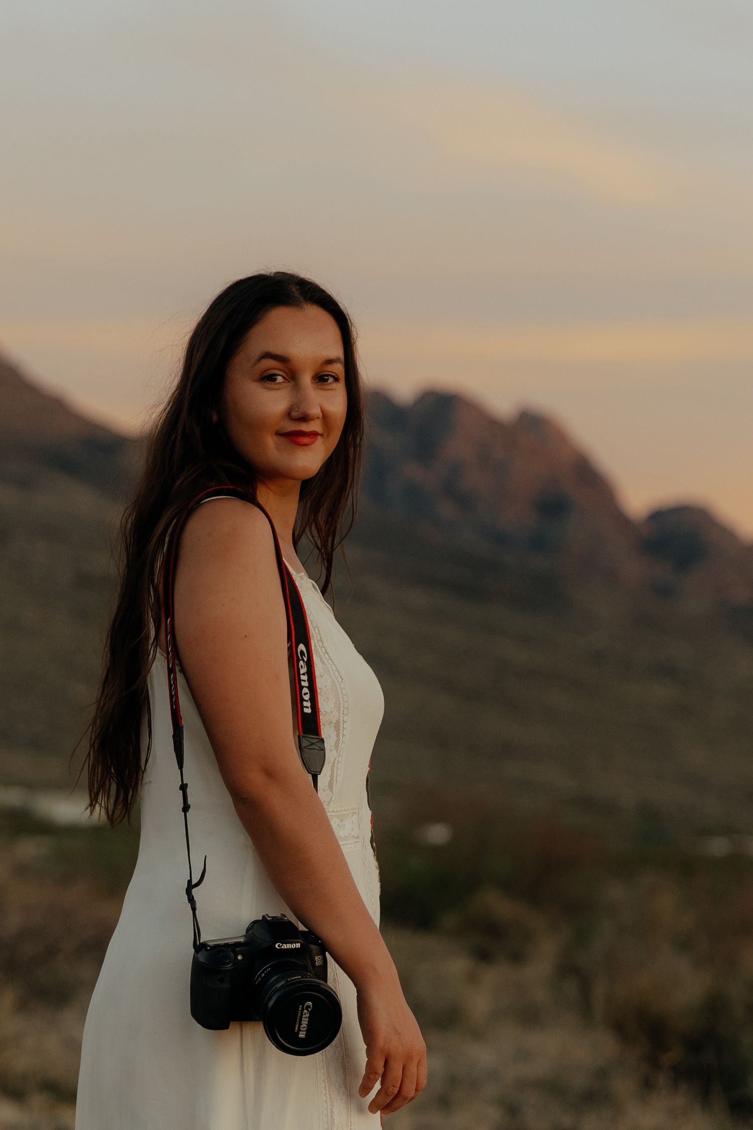 Woman in white dress holding Canon camera poses against mountain sunset backdrop.