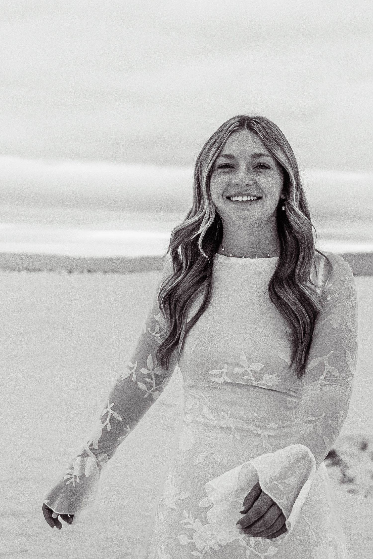 Black and white portrait of smiling young woman in white lace dress on sandy beach.