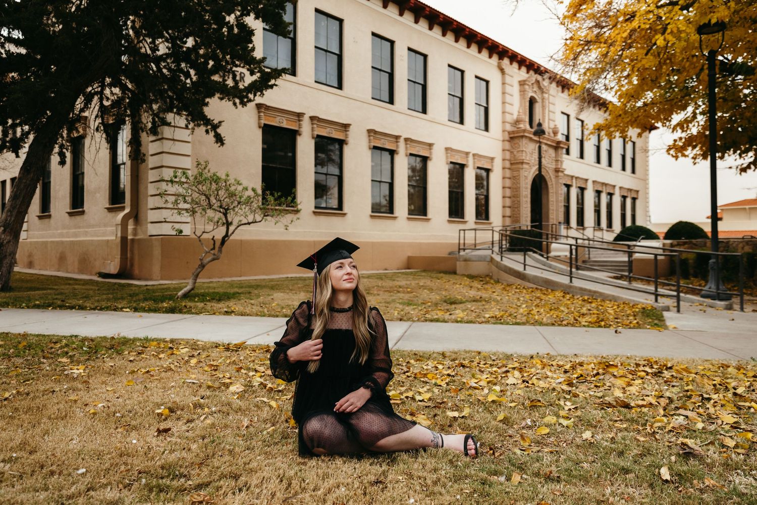 Female graduate in black cap and gown sits on grass outside university building in autumn.