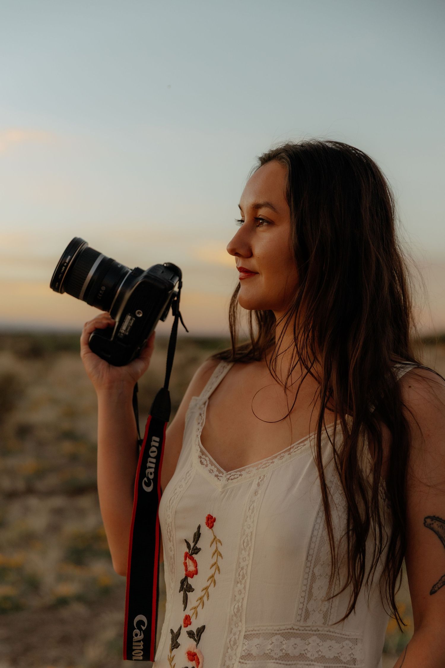 Woman in white embroidered dress holds Canon camera up at desert sunset.