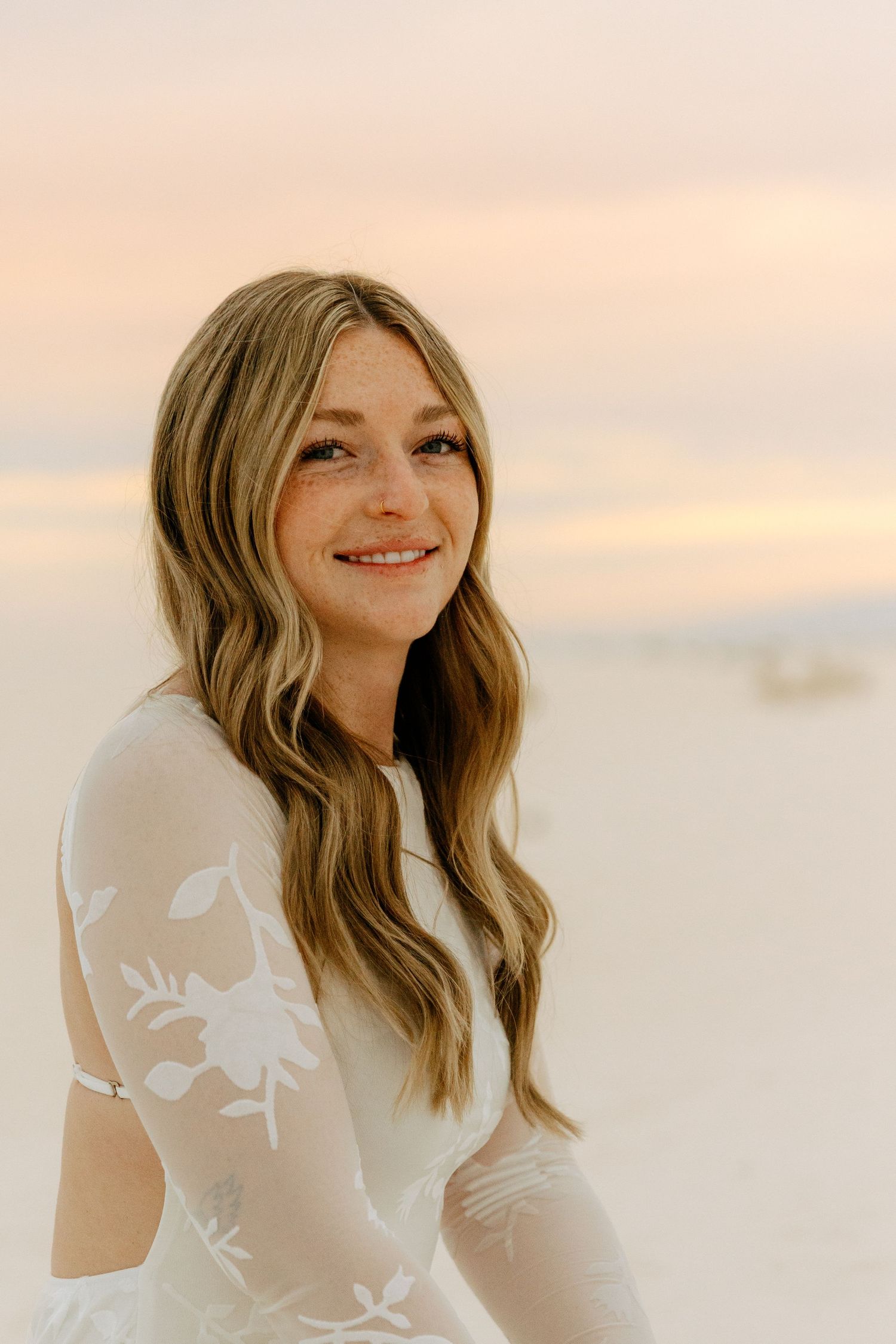 Freckled blonde woman in white floral lace dress smiles warmly while sitting on sandy beach at dusk.