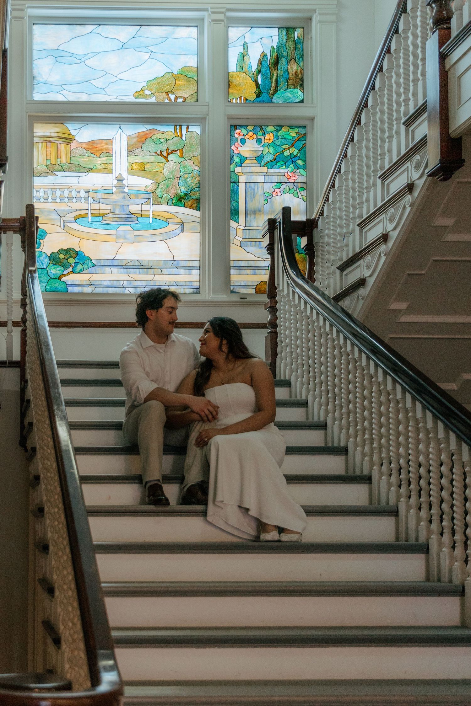Couple sitting together on grand staircase steps with colorful stained glass fountain window behind them.